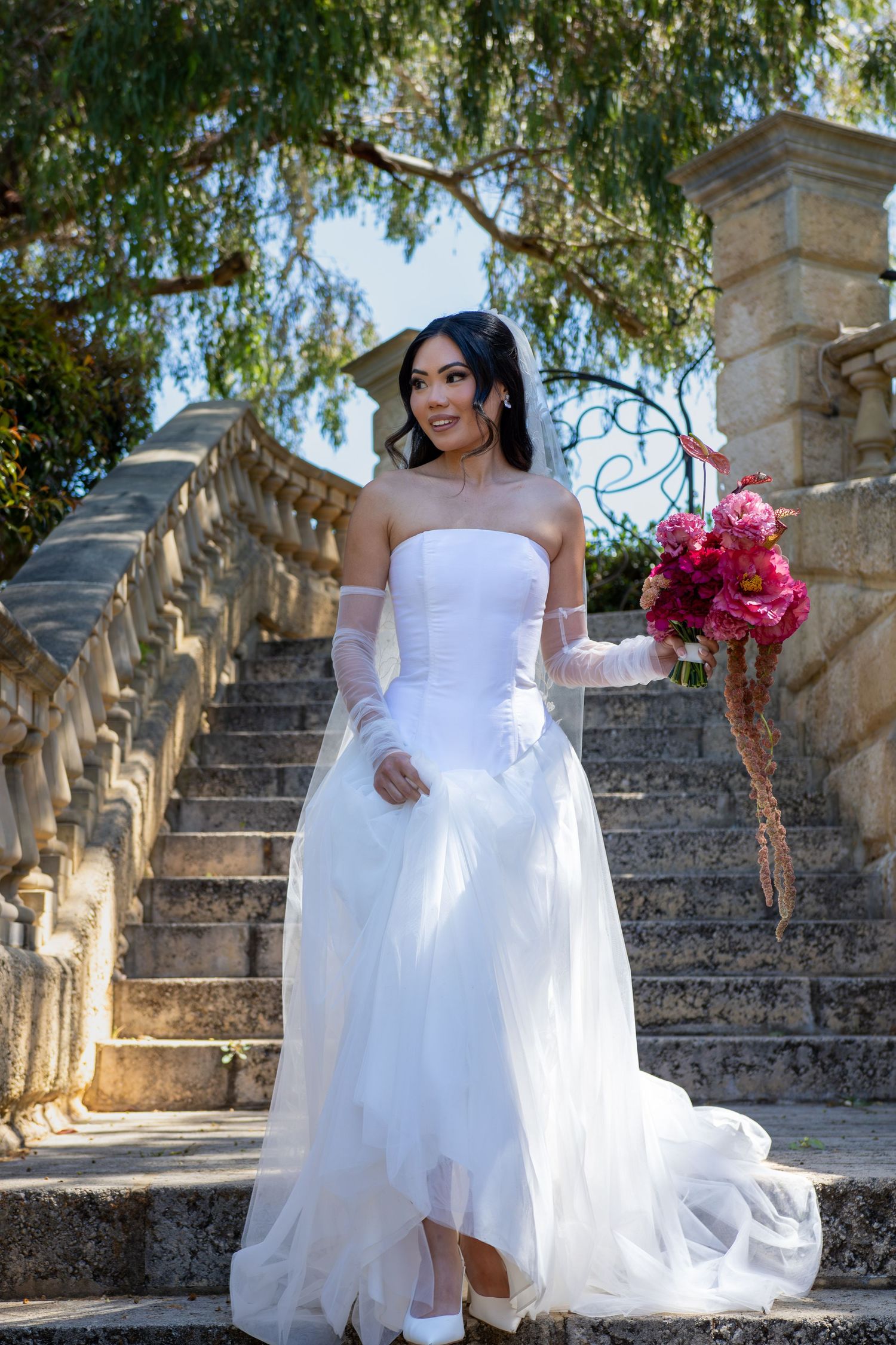 A bride in a flowing white dress poses gracefully on stone steps surrounded by lush greenery and architectural details.