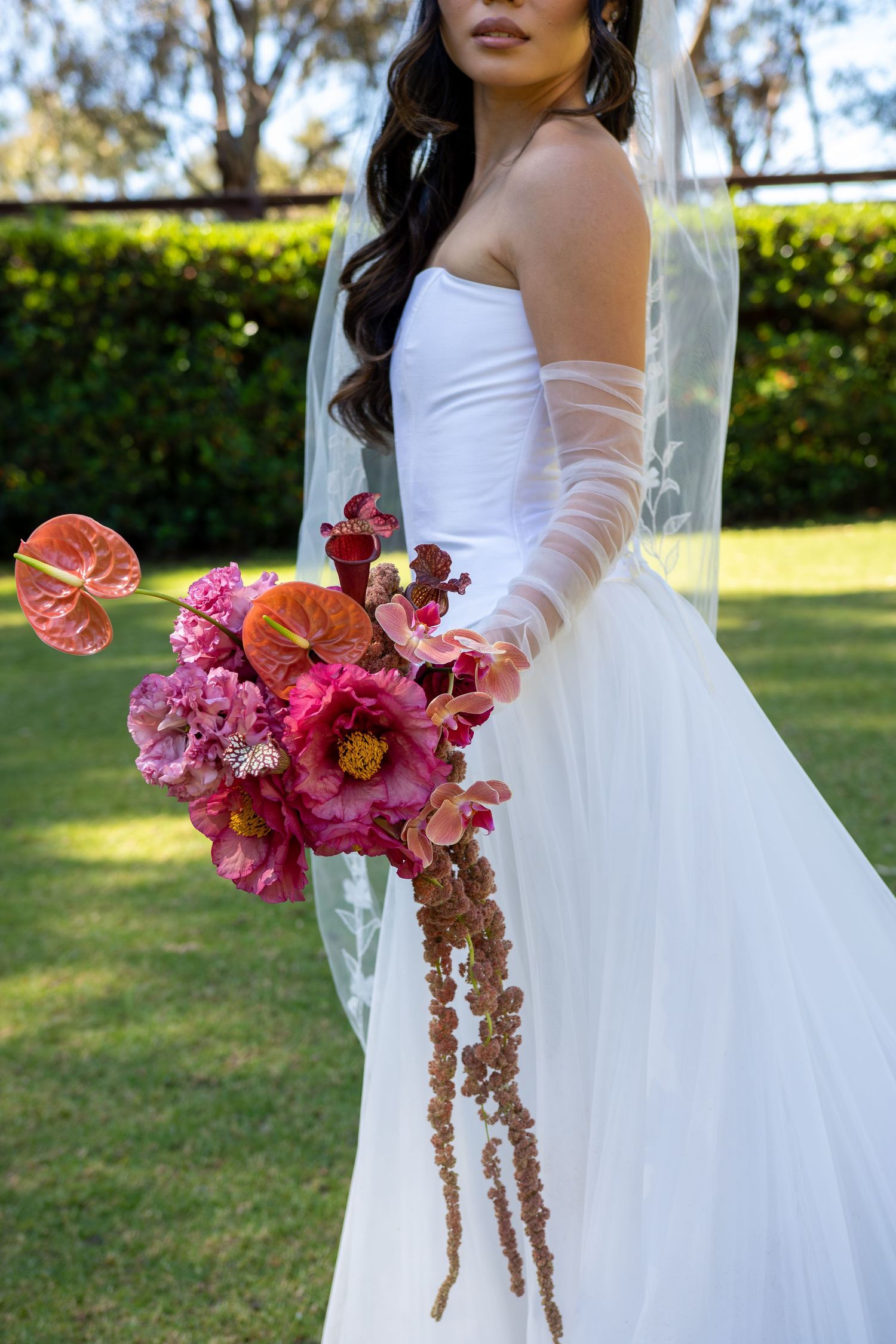 Elegant bridal portrait featuring a white wedding gown with sheer sleeves and a vibrant pink floral bouquet in a garden setting.