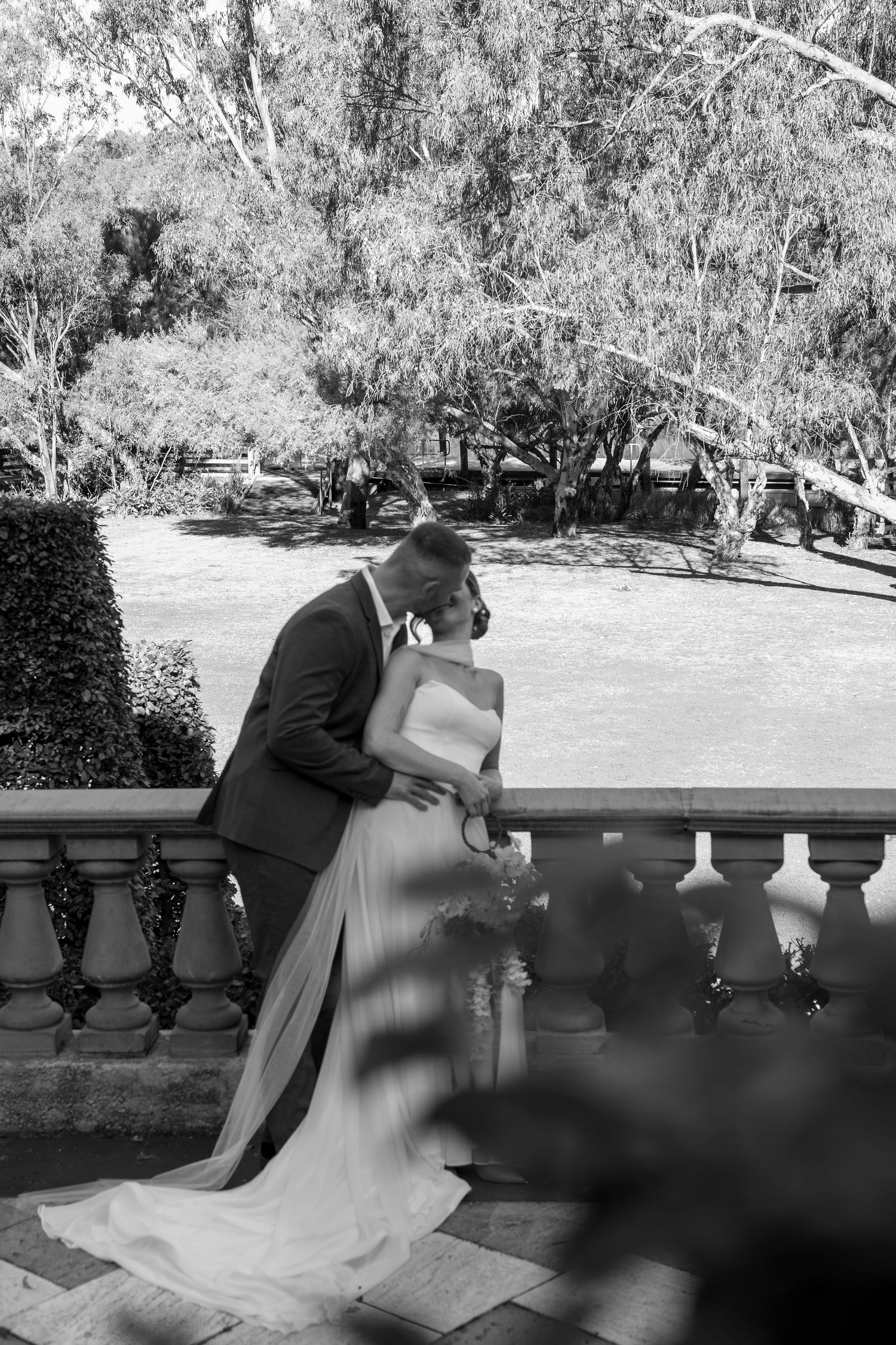 A romantic couple in formal attire shares an intimate moment on a stone balcony in black and white.