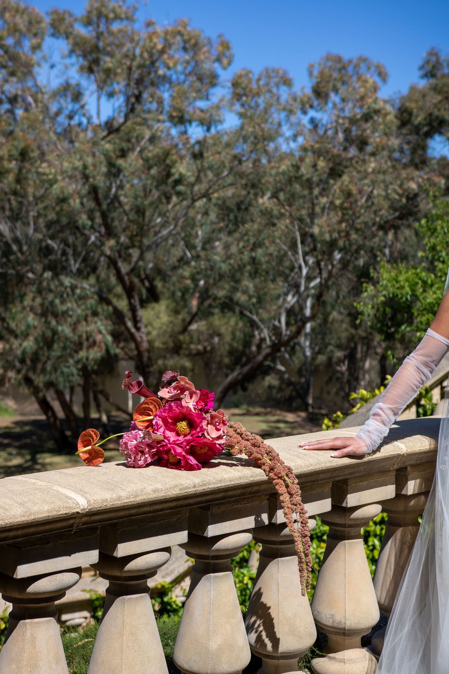 A red and pink floral bouquet rests on a stone balustrade railing with trees in the background on a sunny day.