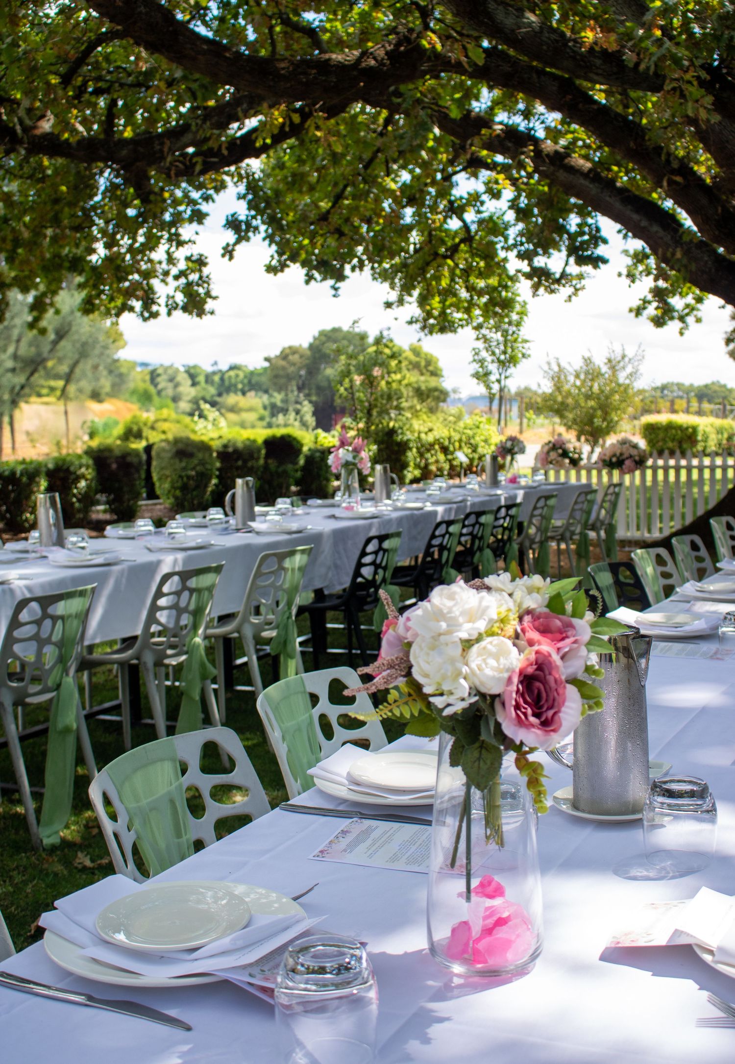 Elegant outdoor wedding reception tables set with white linens, green chairs, and delicate pink floral centerpieces under shady trees.