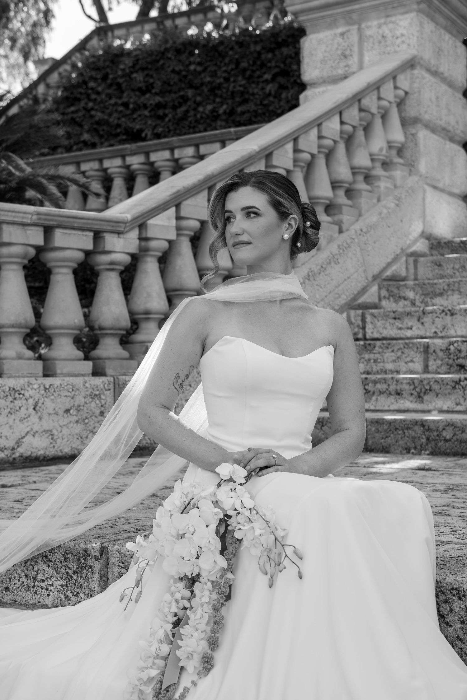 A bride in a white strapless gown poses gracefully on stone steps with a flowing veil and floral bouquet.