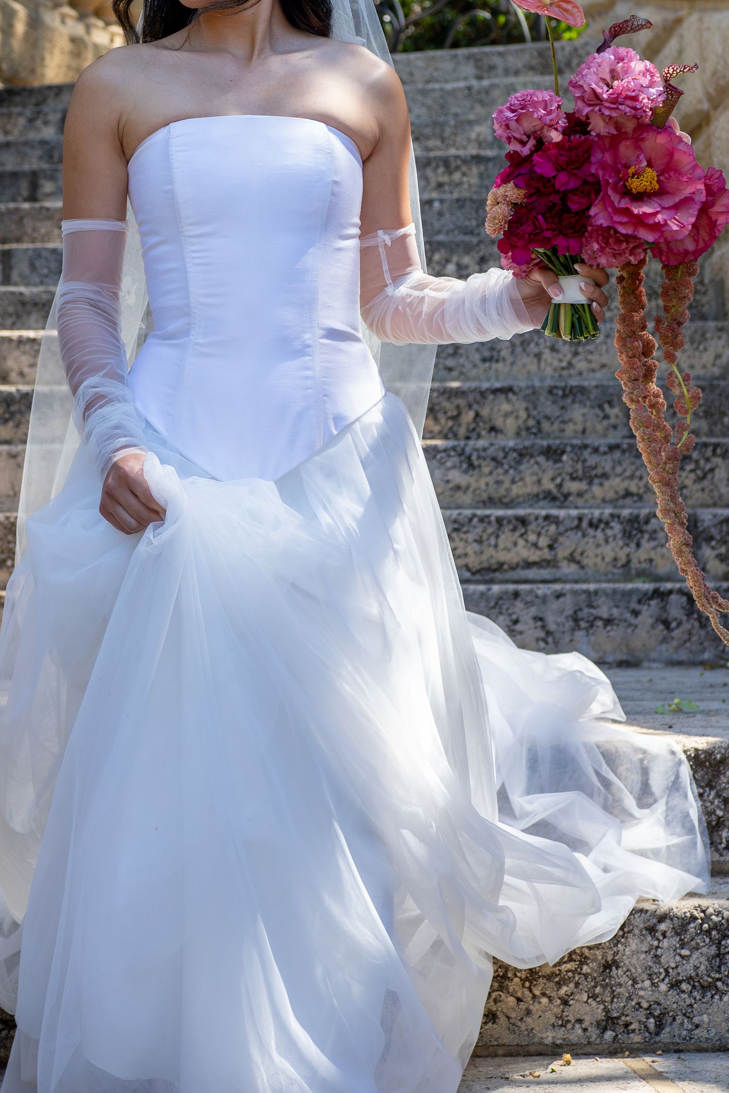 A bride in a flowing white wedding dress walks down stone steps while holding a vibrant pink bouquet.