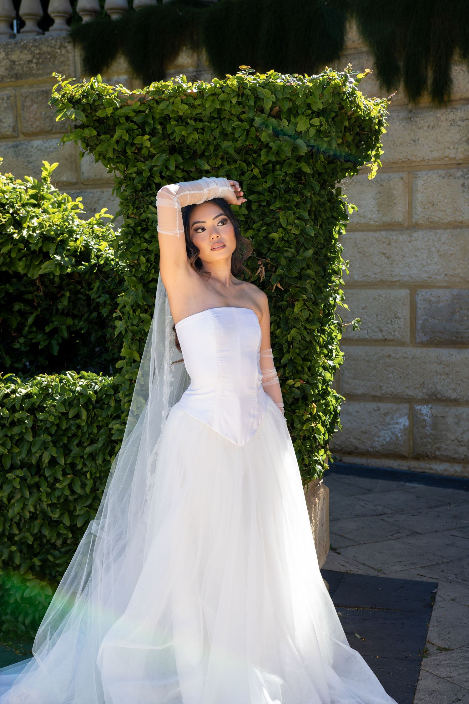 A bride in a strapless white wedding dress poses playfully with her veil in front of lush garden greenery.