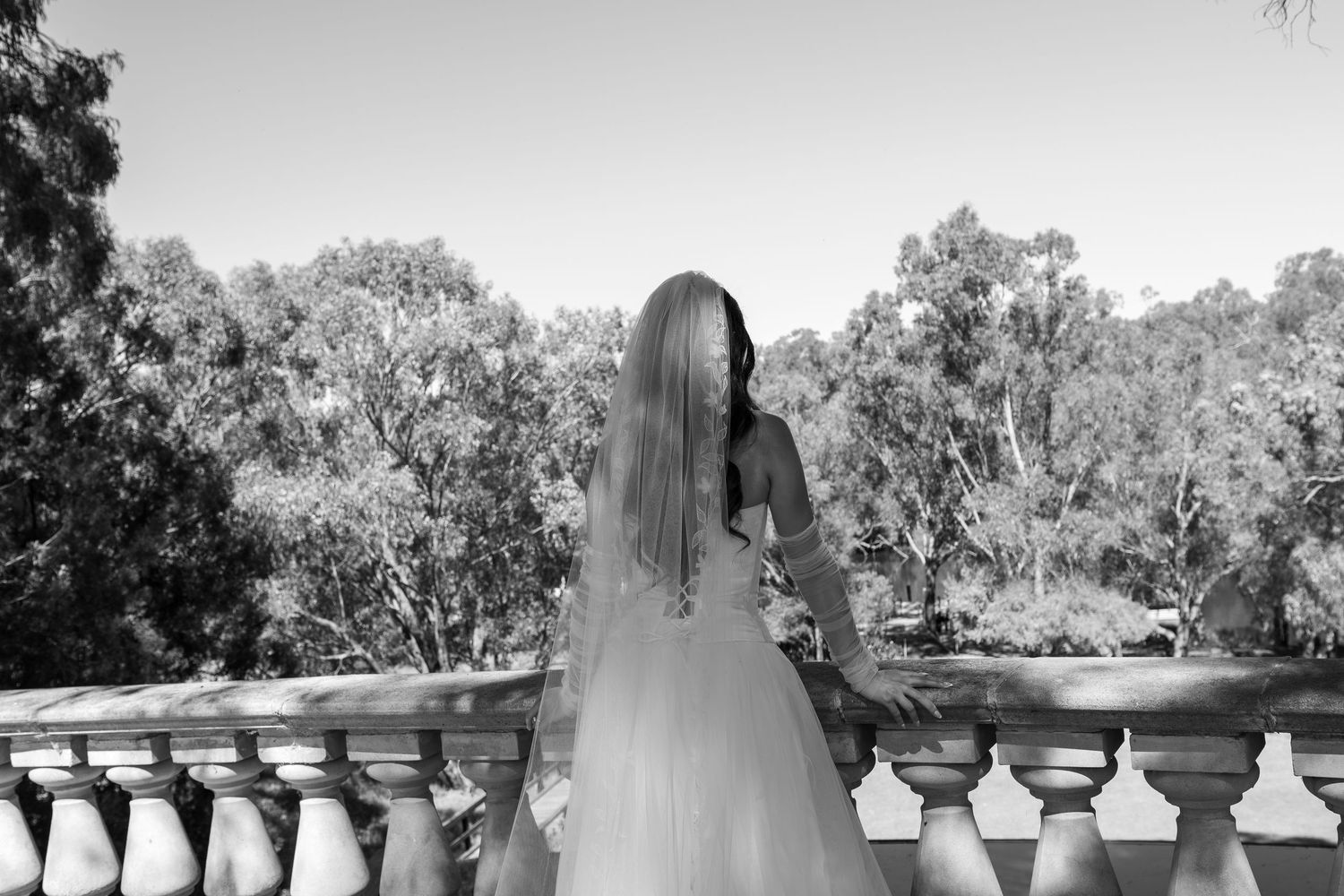 A bride in a white wedding dress stands at a stone balustrade overlooking trees in black and white photography.