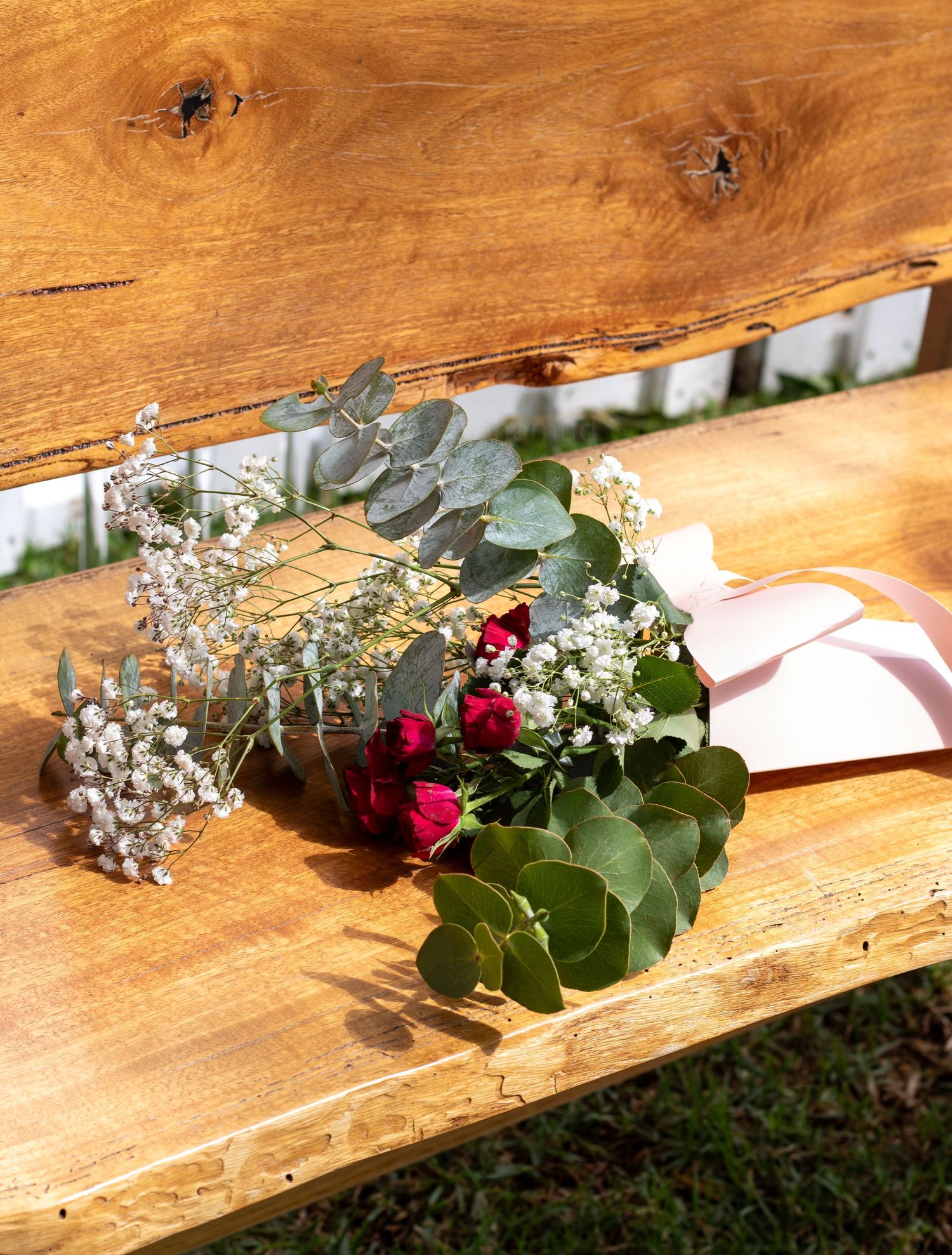A rustic wooden bench decorated with a floral arrangement of red roses, baby's breath, and eucalyptus leaves.