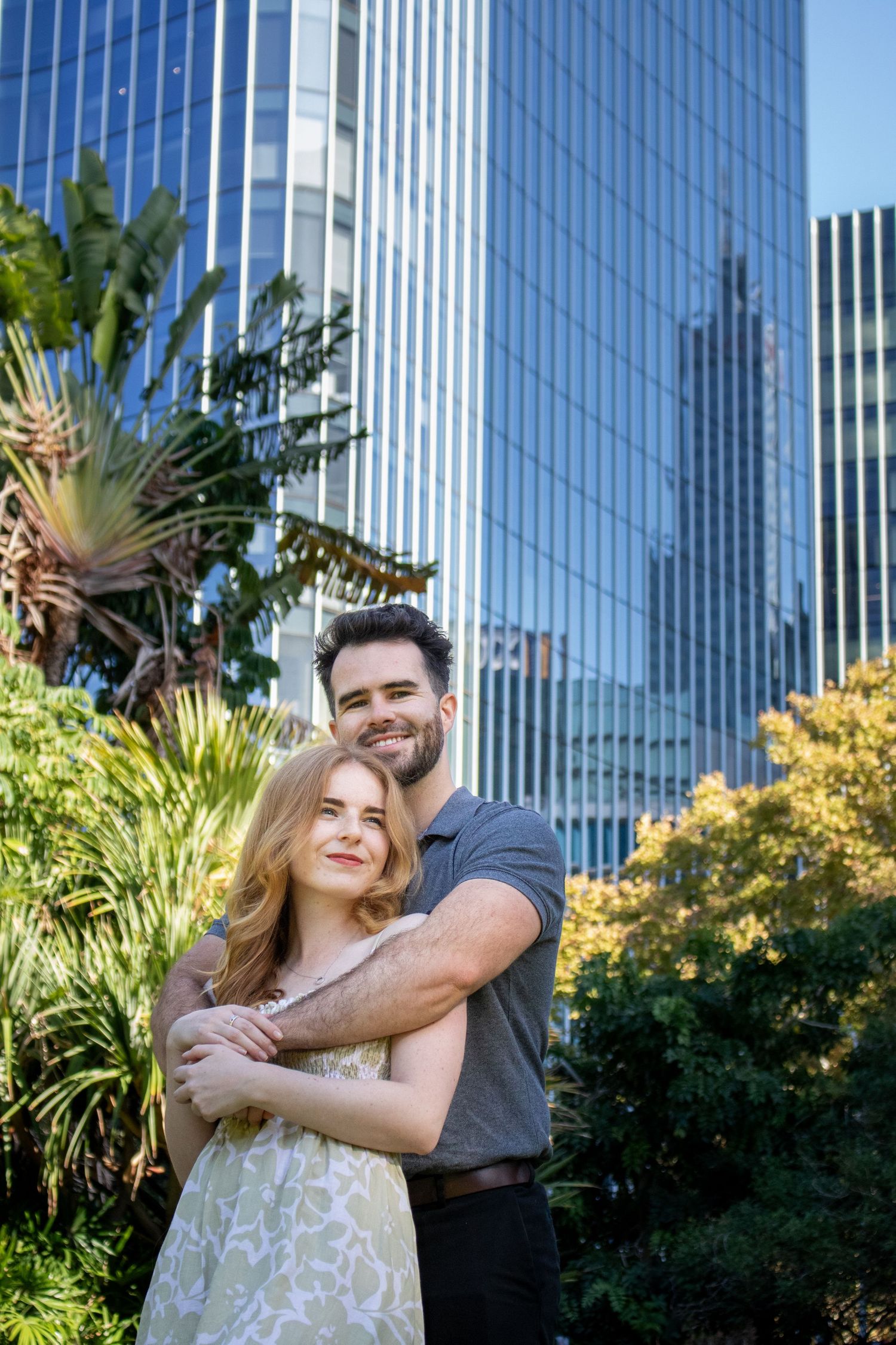 Romantic couple portrait with modern city architecture and lush garden background.