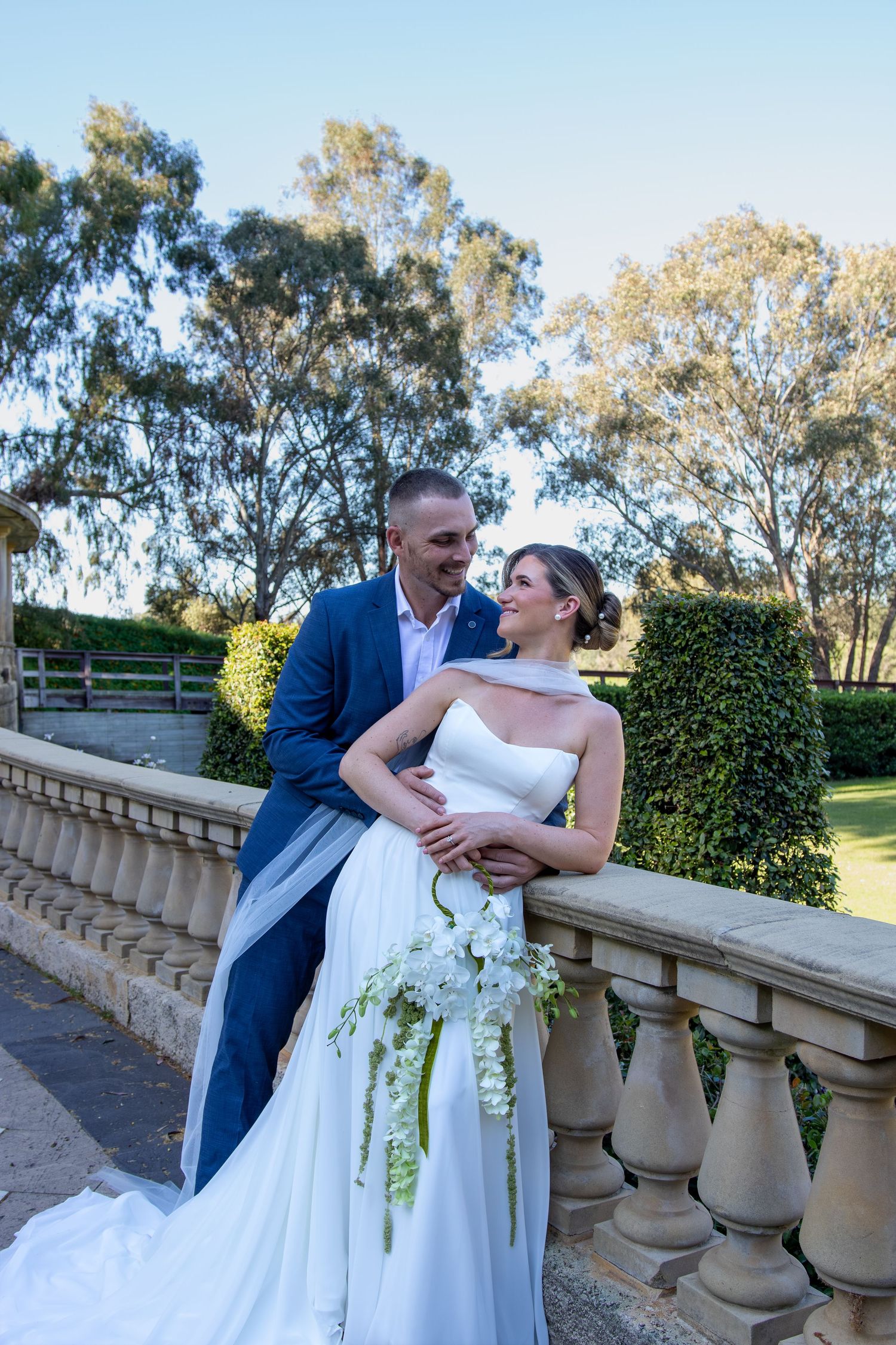 A couple in wedding attire embrace on an elegant stone balcony with lush gardens and trees in the background.