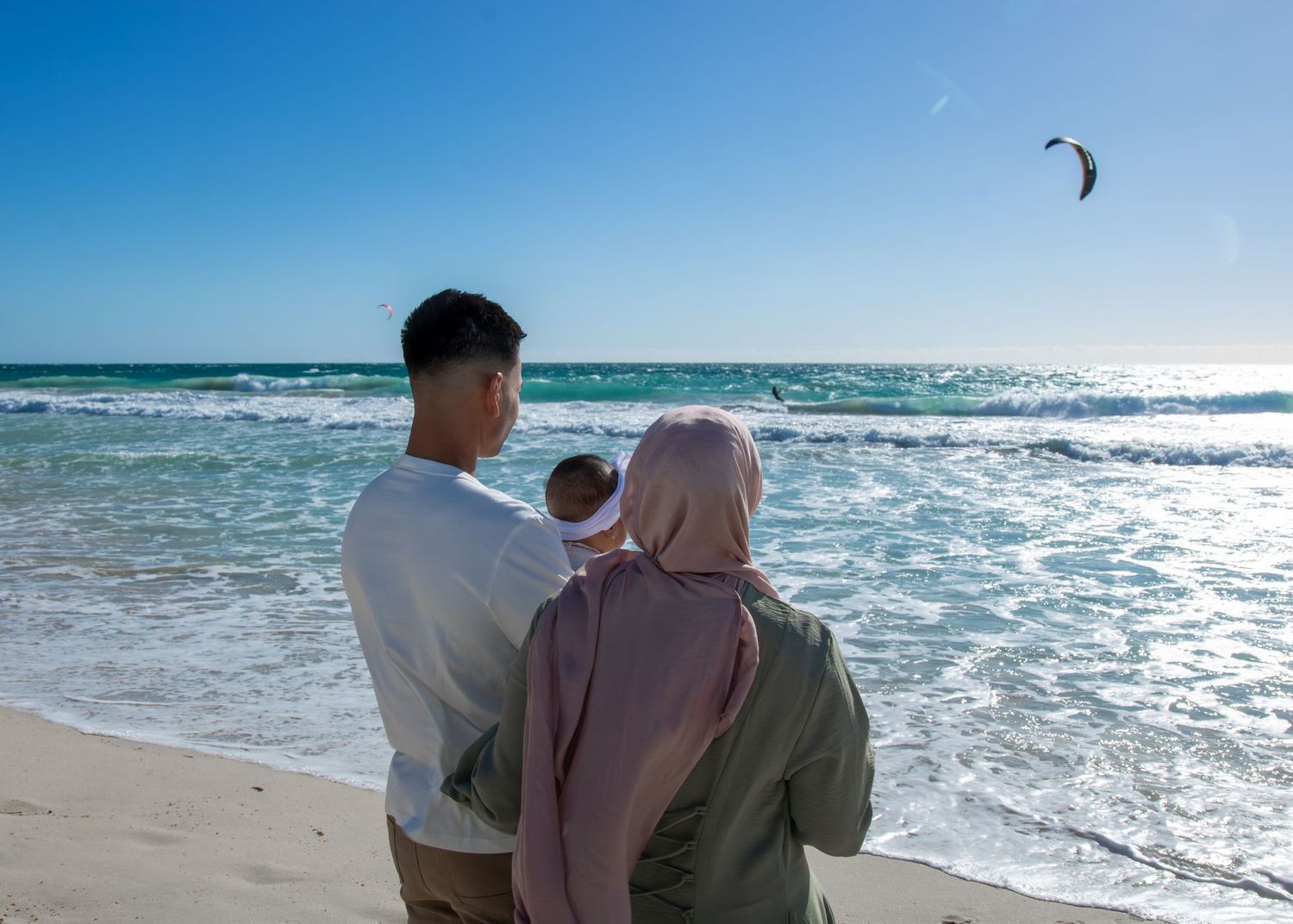 A Muslim couple with a baby enjoys a serene beach view, waves gently lapping the shore under a clear blue sky.