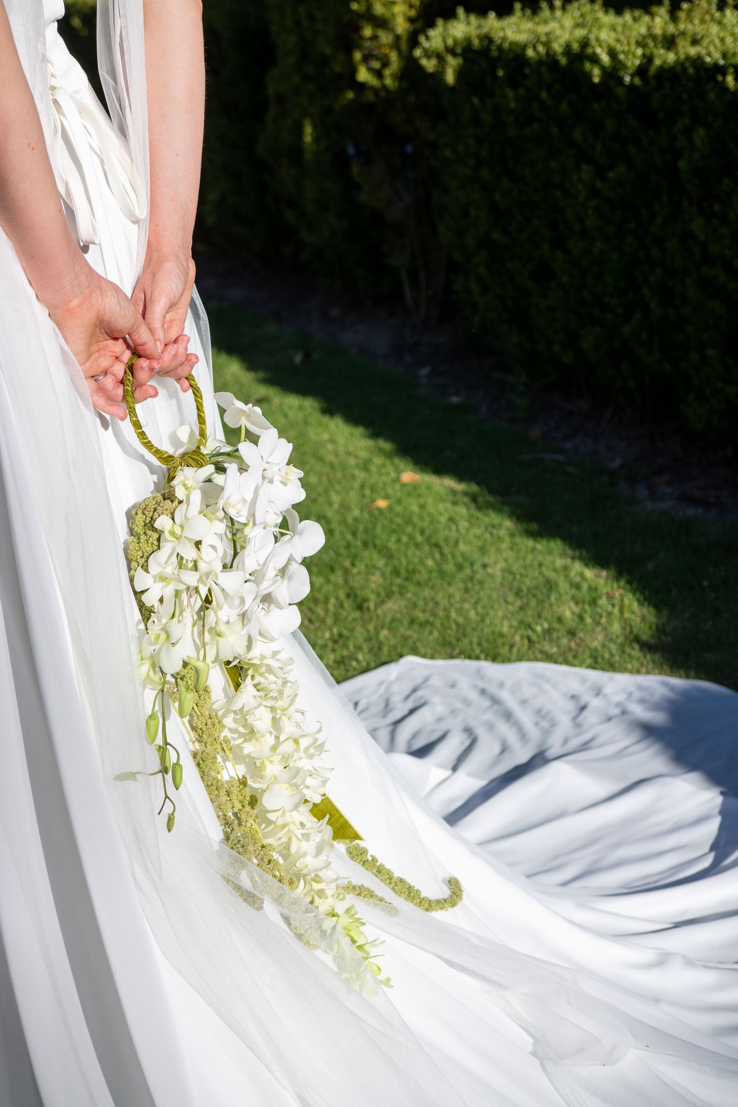 Close-up view of a cascading white orchid bouquet against a flowing white wedding dress in an outdoor garden setting.