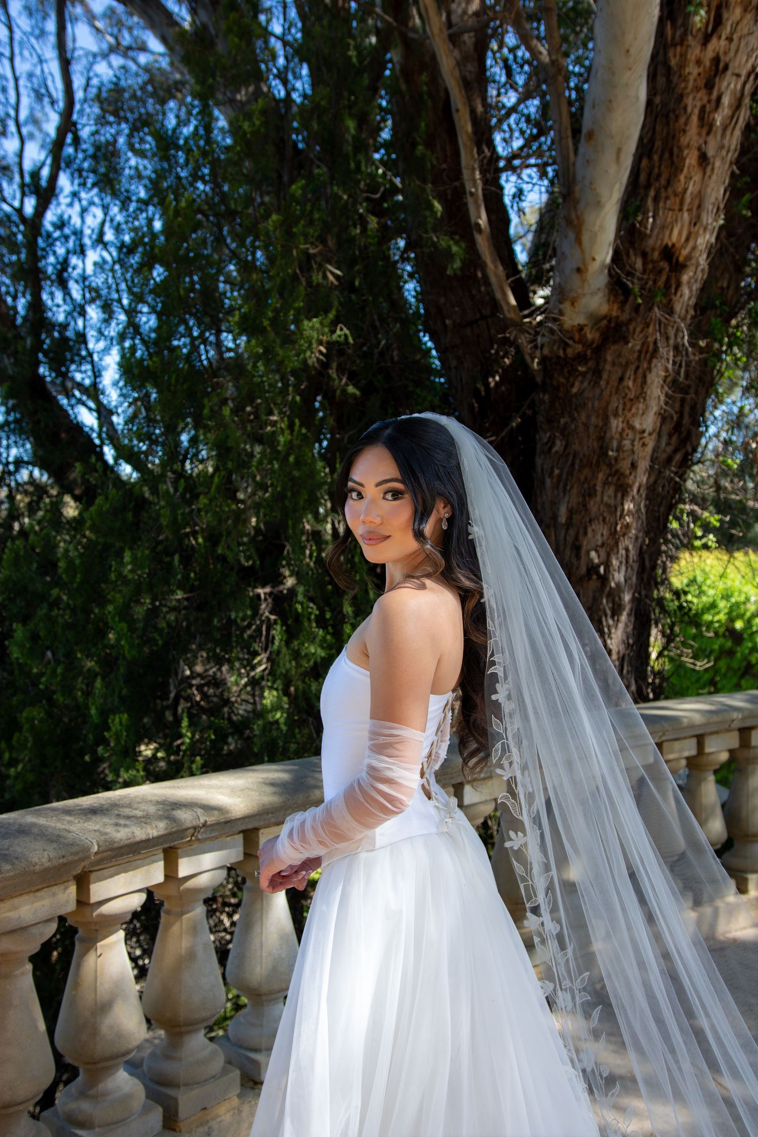A bride in a white wedding dress poses gracefully against a backdrop of trees and stone balustrades.