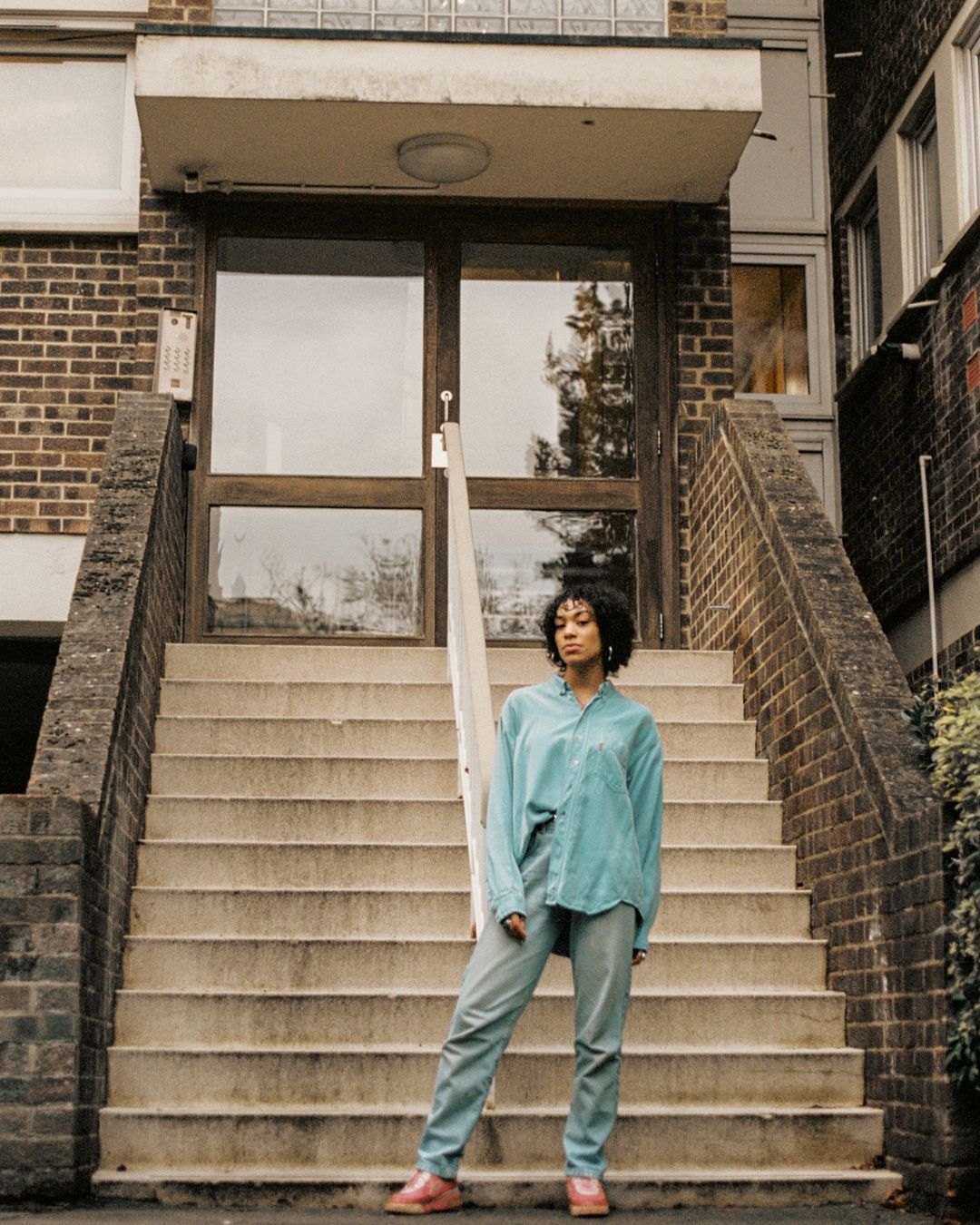 Model in mint shirt and jeans poses on concrete steps of brick building entrance.