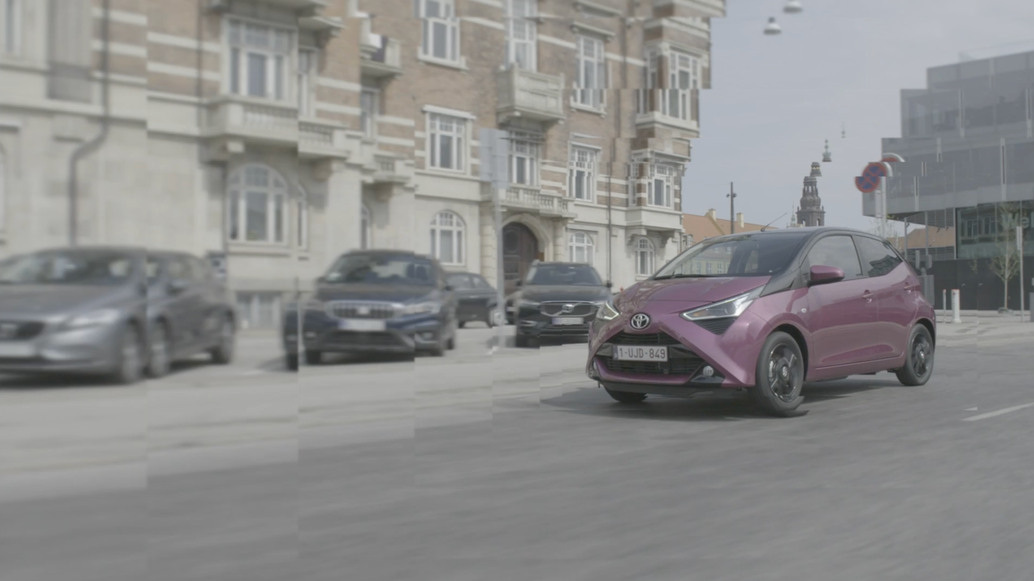 Pink car driving on a city street with other parked vehicles and buildings in the background.