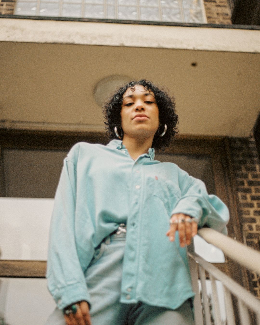 Candid moment in oversized mint shirt leaning on stair railing of building entrance.