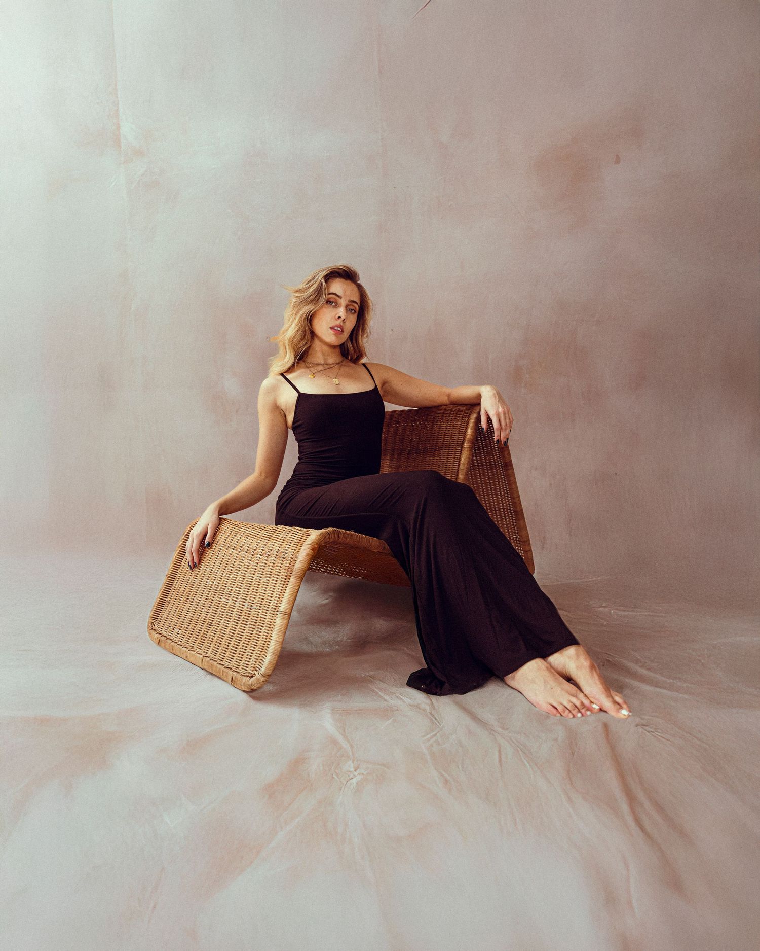 Elegant pose in black outfit seated on wicker furniture against neutral backdrop.