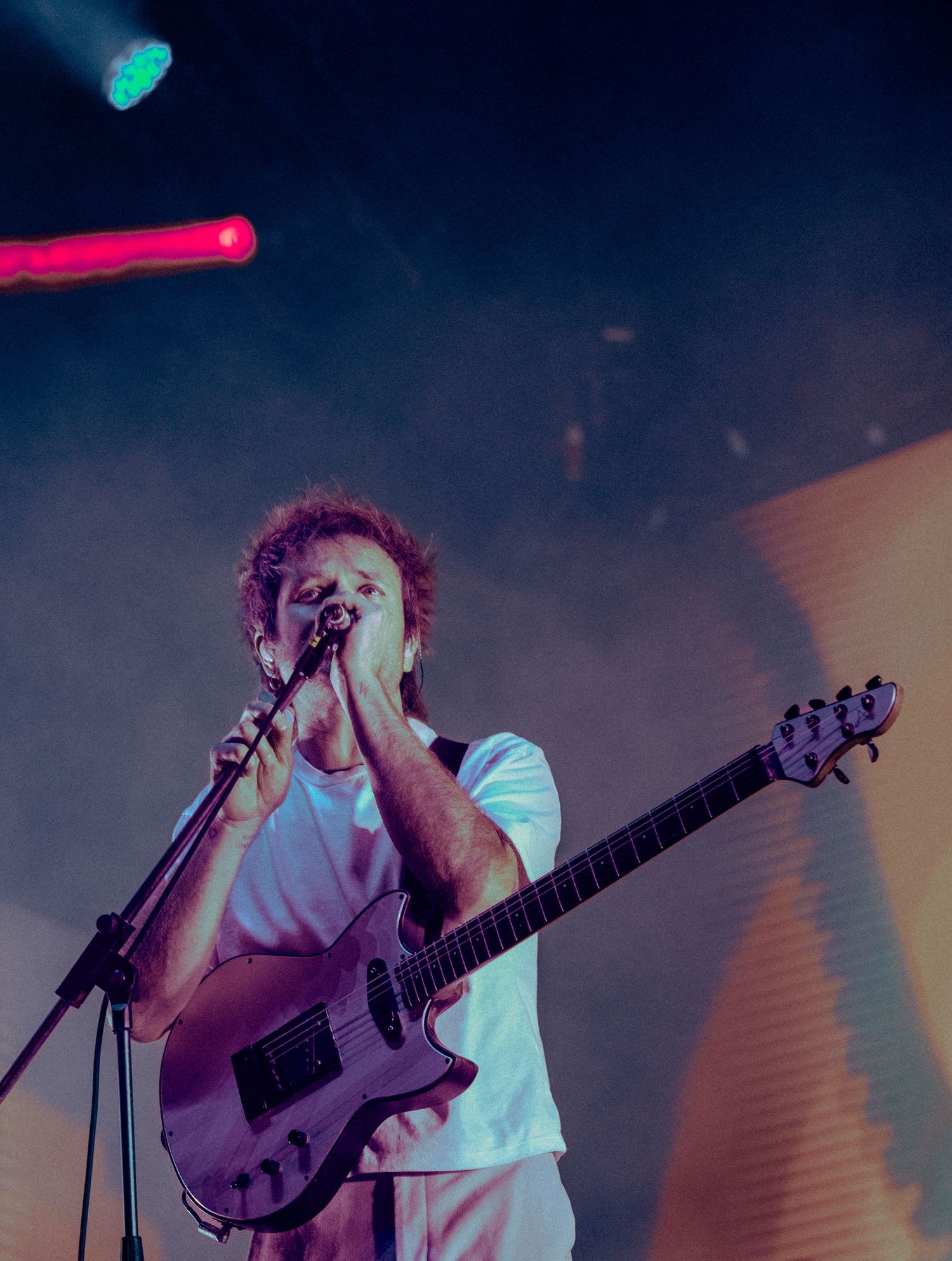 Musician performing on stage under blue and pink lighting with guitar and microphone in atmospheric concert setting.