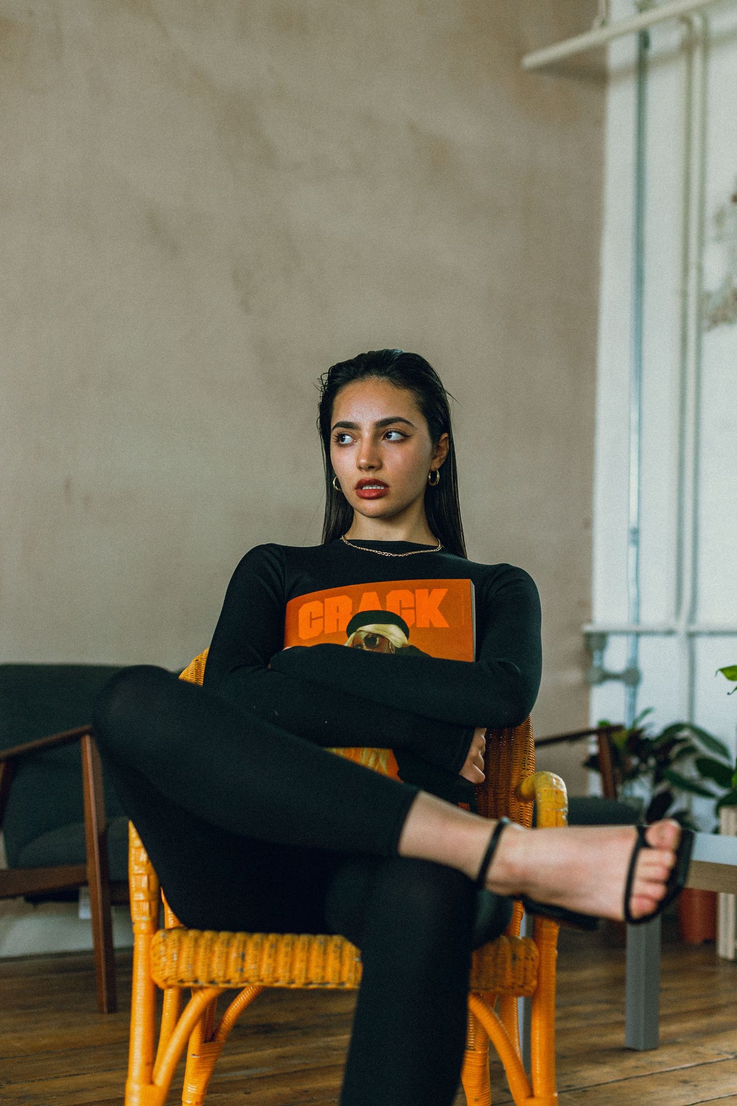 Modern fashion shot in black outfit against neutral wall with rattan furniture.
