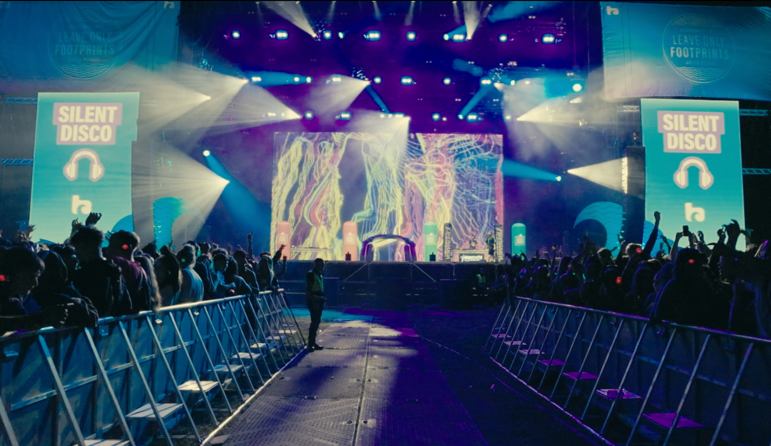 Concert venue with colorful stage lighting, metal barriers, and large video screens displaying performance content.
