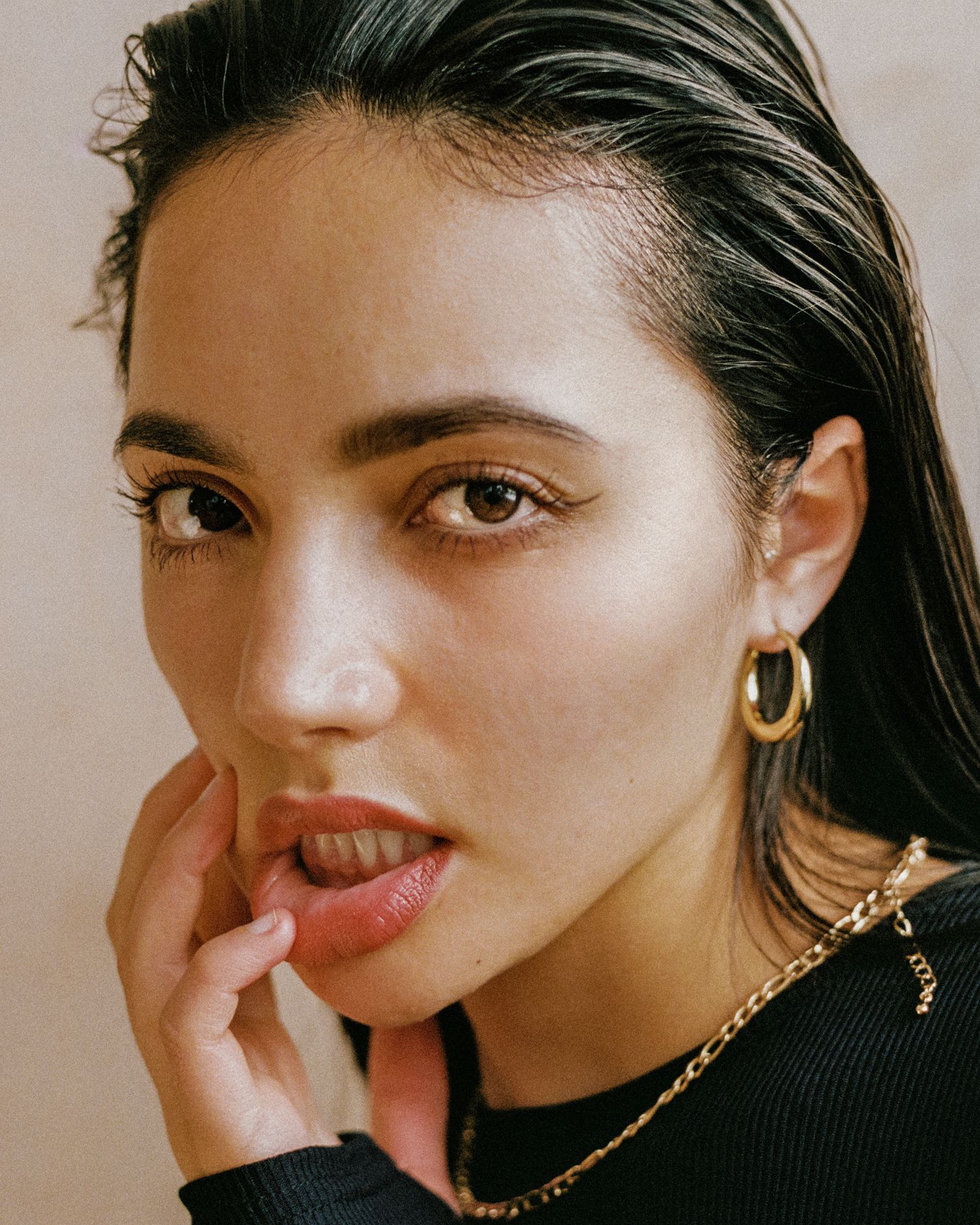 A close-up beauty portrait showing glossy lips and gold hoop earrings against a neutral background.