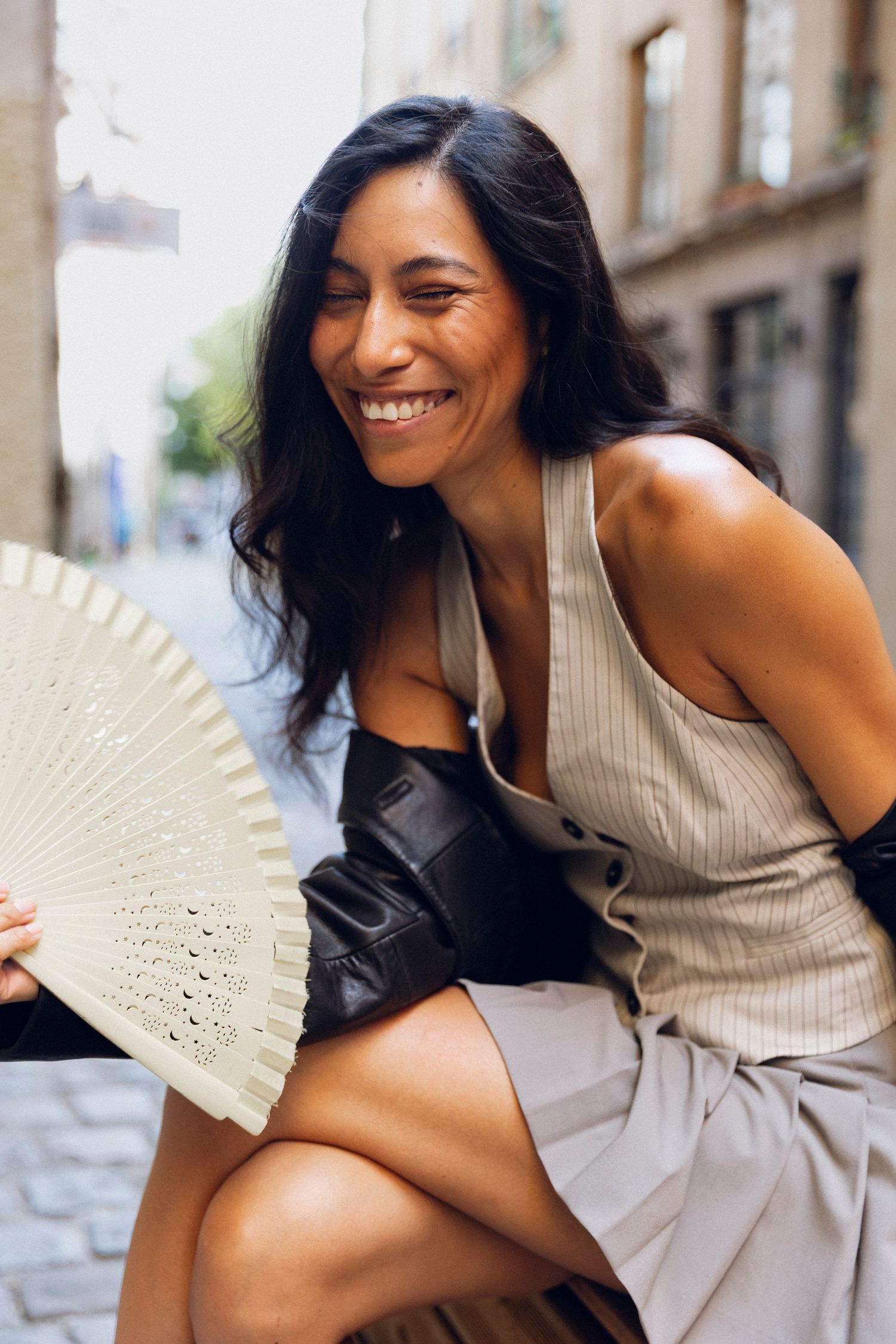 Fashion model in beige sleeveless top poses playfully with decorative fan against urban backdrop.