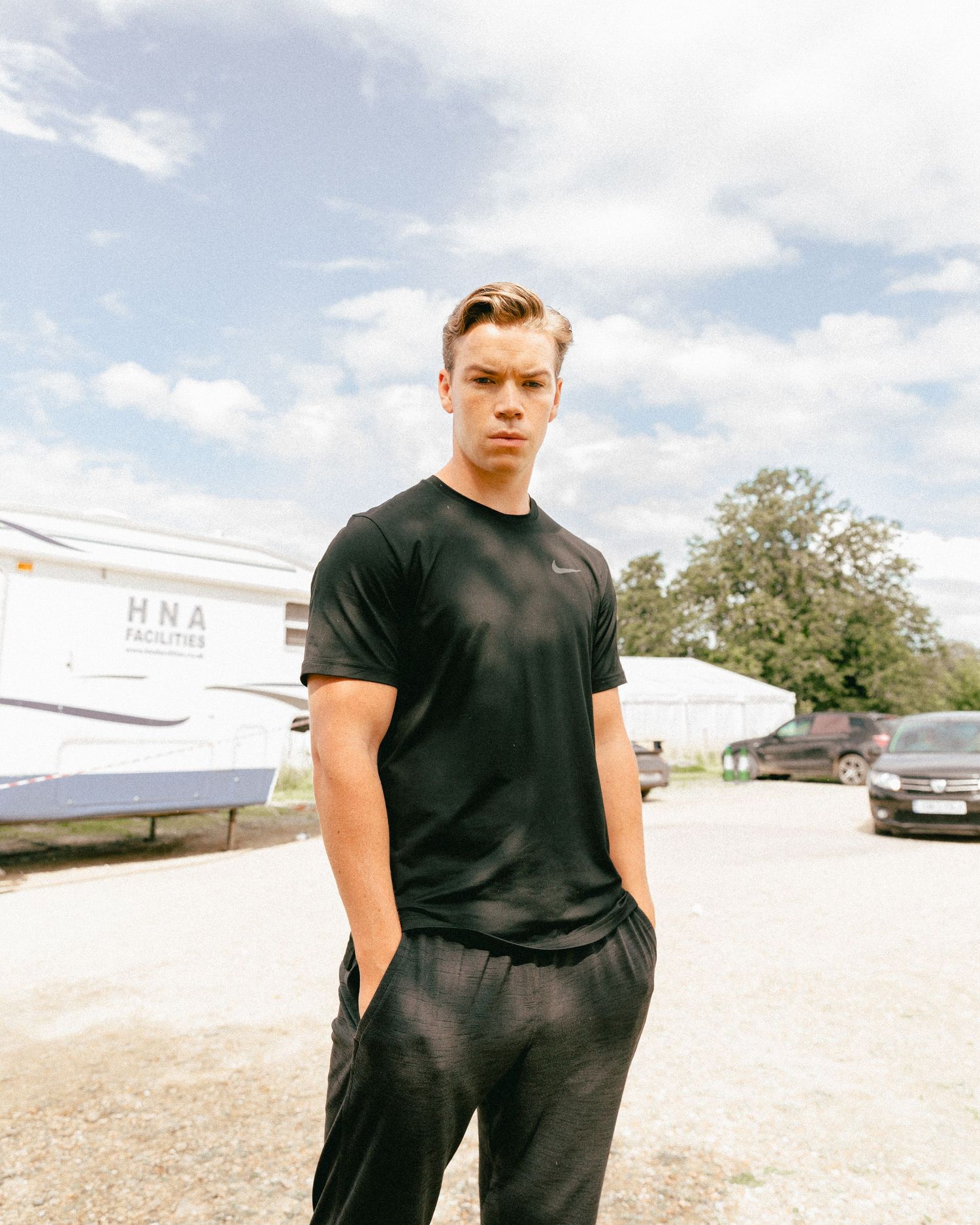 A young person wearing a black t-shirt poses outdoors against a cloudy sky.