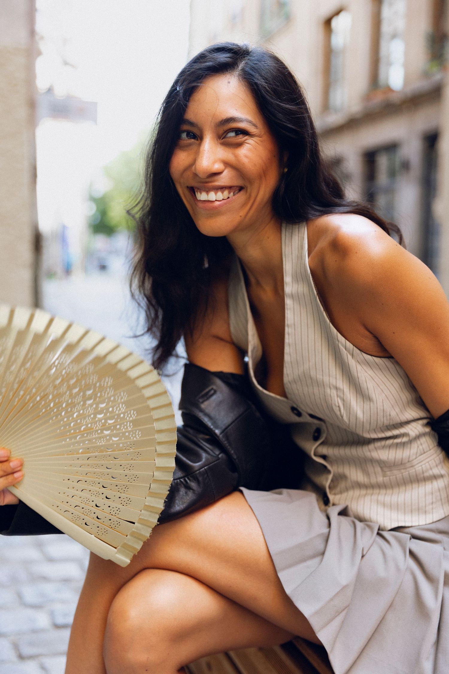 Fashion model in beige sleeveless top poses playfully with decorative fan against urban backdrop.