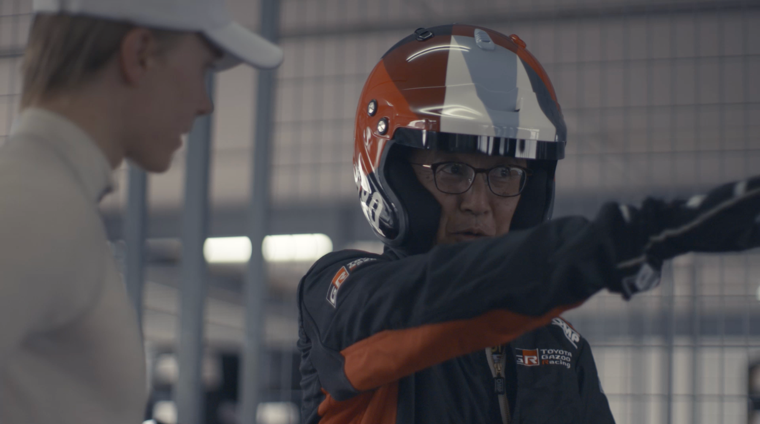 Two people in motorsport gear, one pointing outward while wearing a racing helmet.