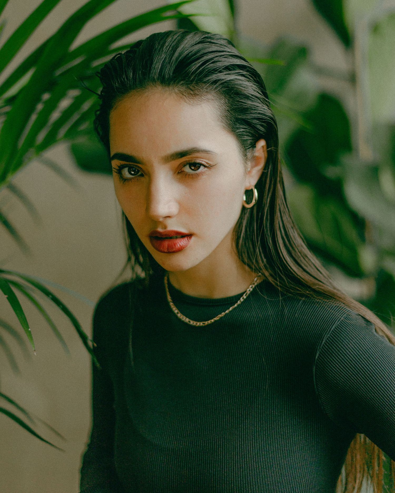 Elegant portrait with palm fronds and gold chain necklace against neutral wall.