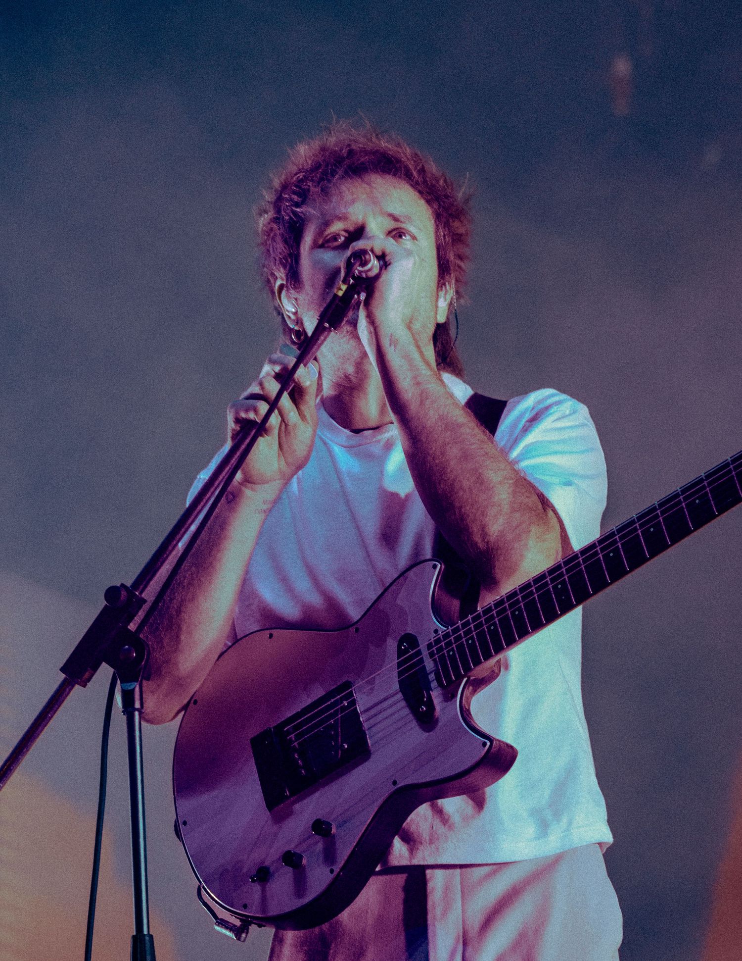 A performer sings passionately into a microphone while playing bass guitar on a dimly lit concert stage.