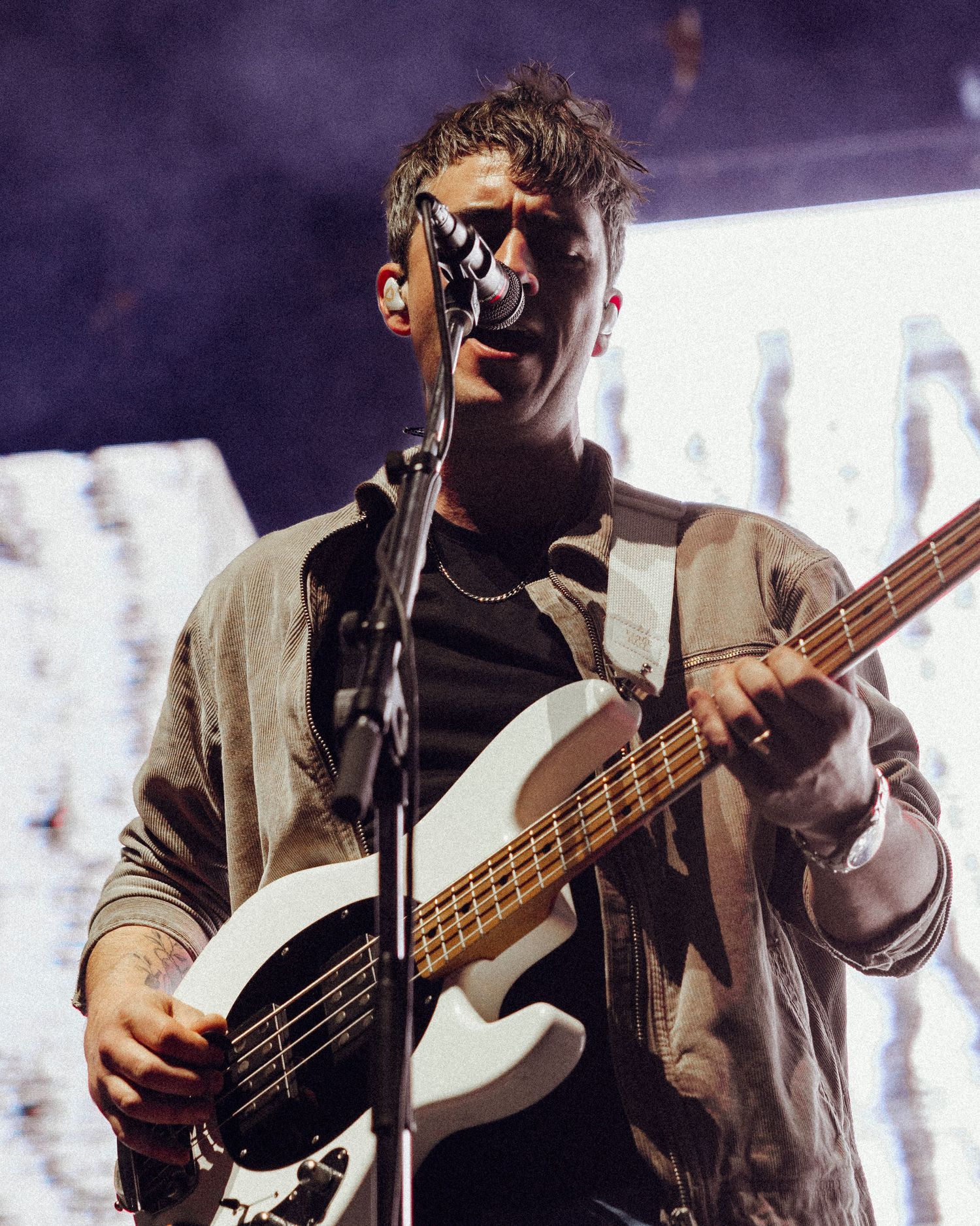 Musician performs on stage with a white bass guitar while singing into microphone with atmospheric lighting and smoke effects.