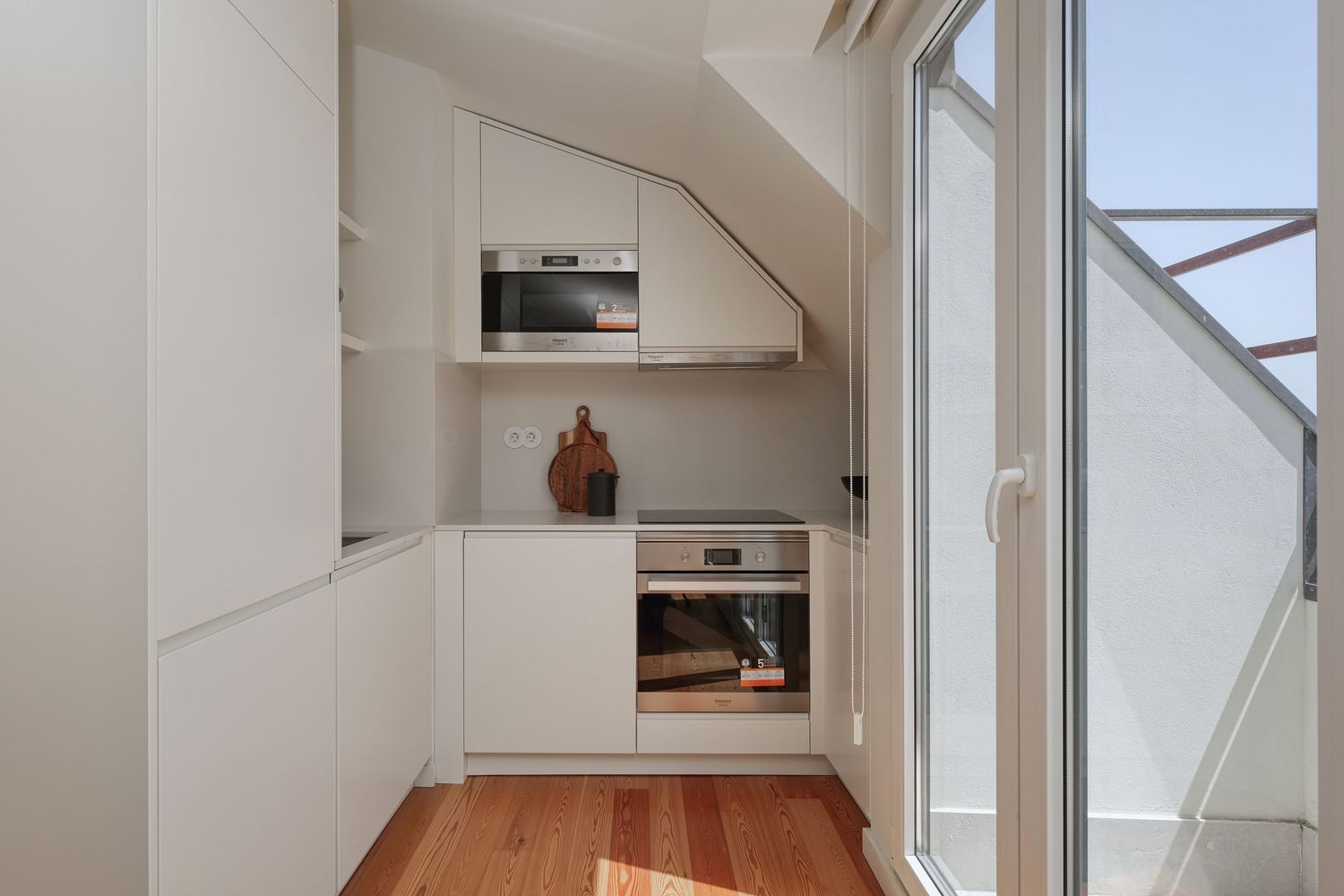 Compact white kitchen under sloped ceiling with built-in appliances, wood floors, and glass balcony door.