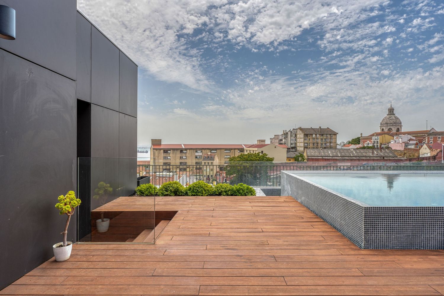 Modern rooftop terrace with wooden deck, infinity pool, and panoramic city views under a partly cloudy sky.