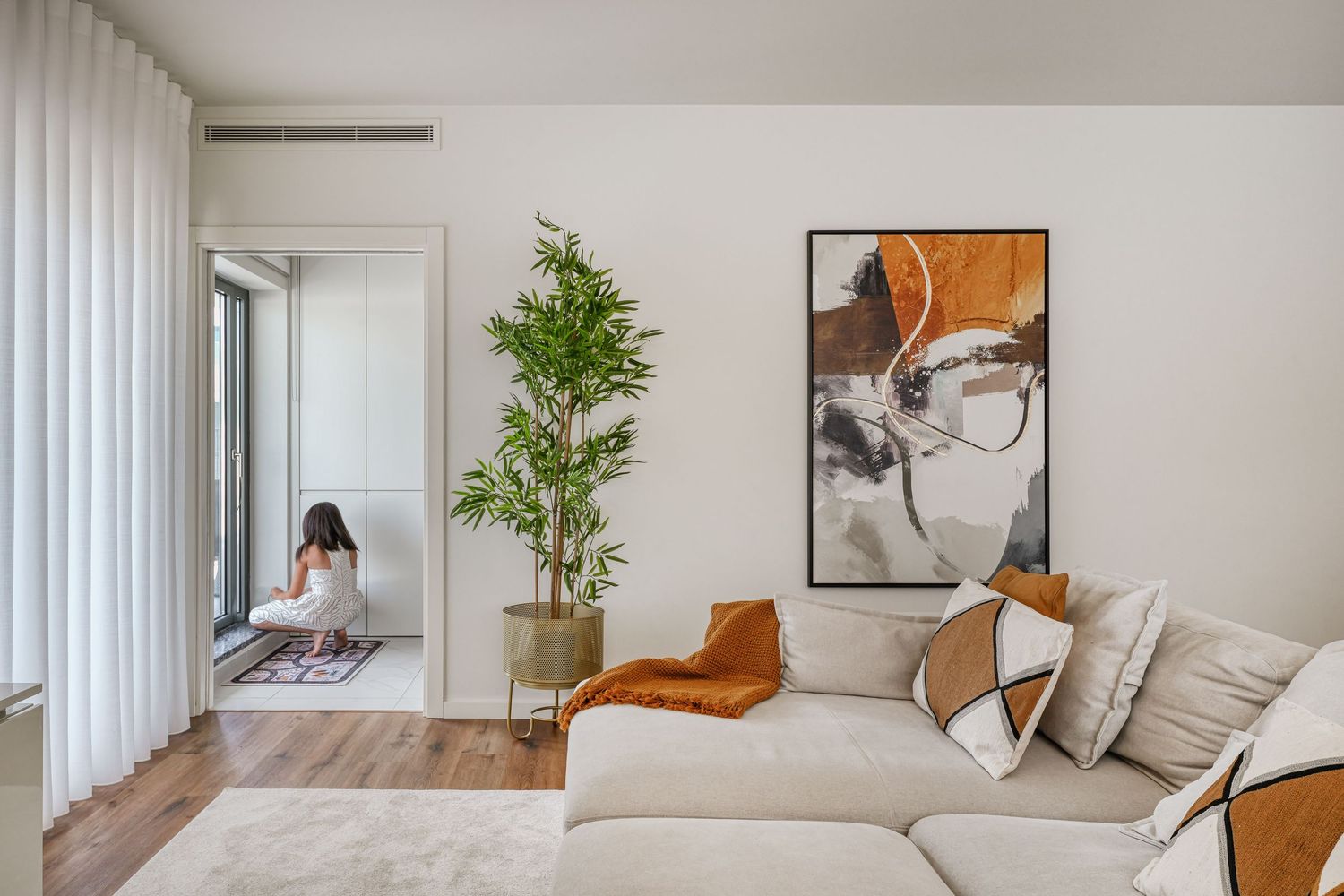 Modern living room with beige sectional sofa, abstract art, potted bamboo plant, and sheer curtains with natural light.