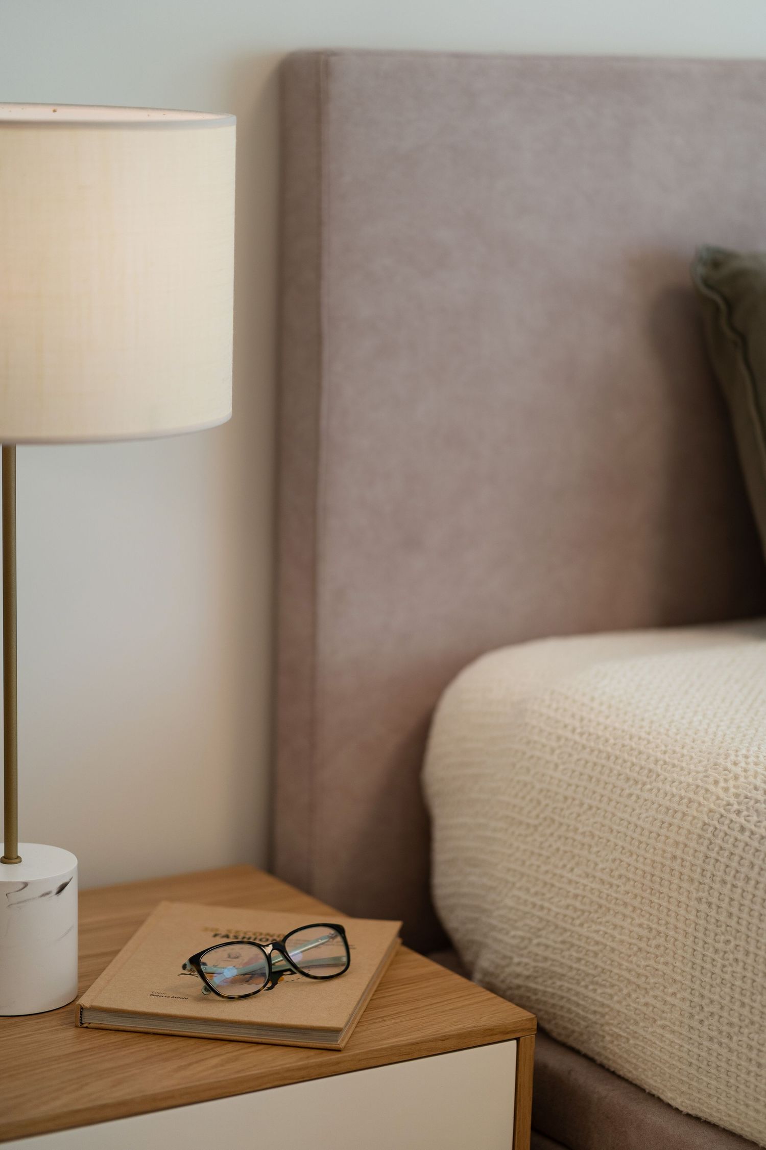 Cozy bedroom nightstand with a marble base lamp, reading glasses, and book beside an upholstered beige headboard.