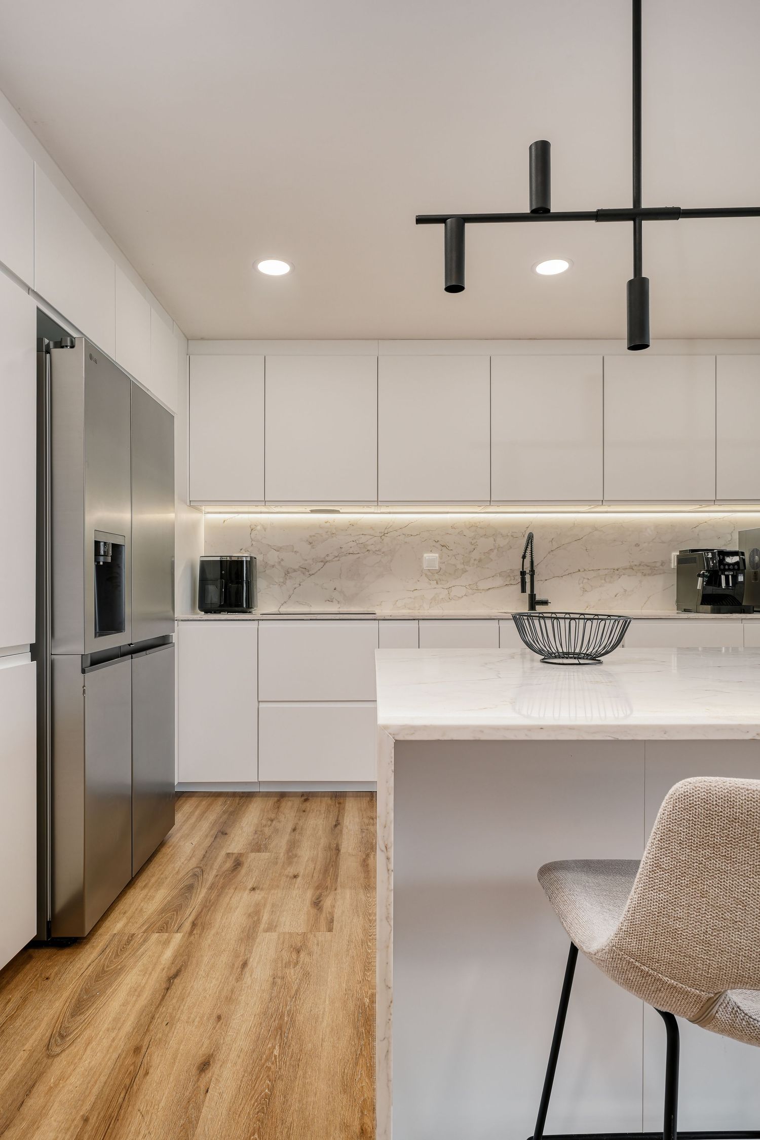 Modern white kitchen with marble backsplash, stainless steel fridge, island seating, black pendant lights, and light wood floors.