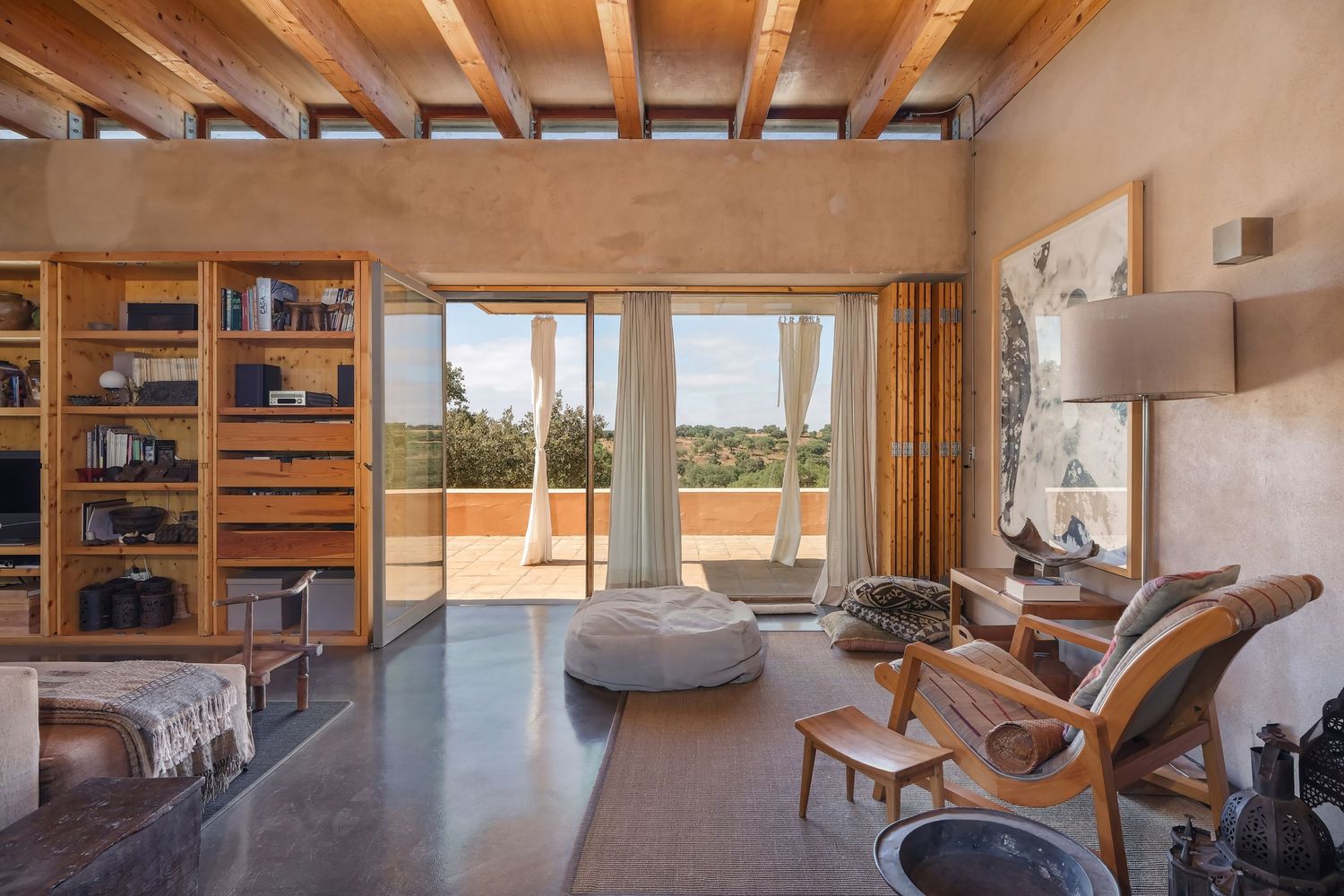 Sunlit living room with wooden beams, built-in bookshelves, and panoramic windows overlooking nature.