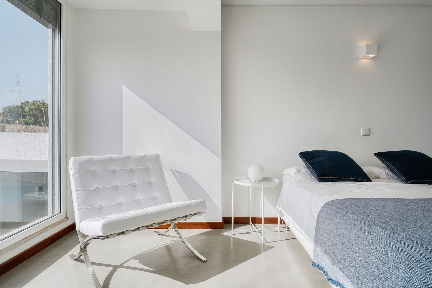 Modern white bedroom with Barcelona chair, large window, blue accent pillows, and minimalist decor in bright natural light.