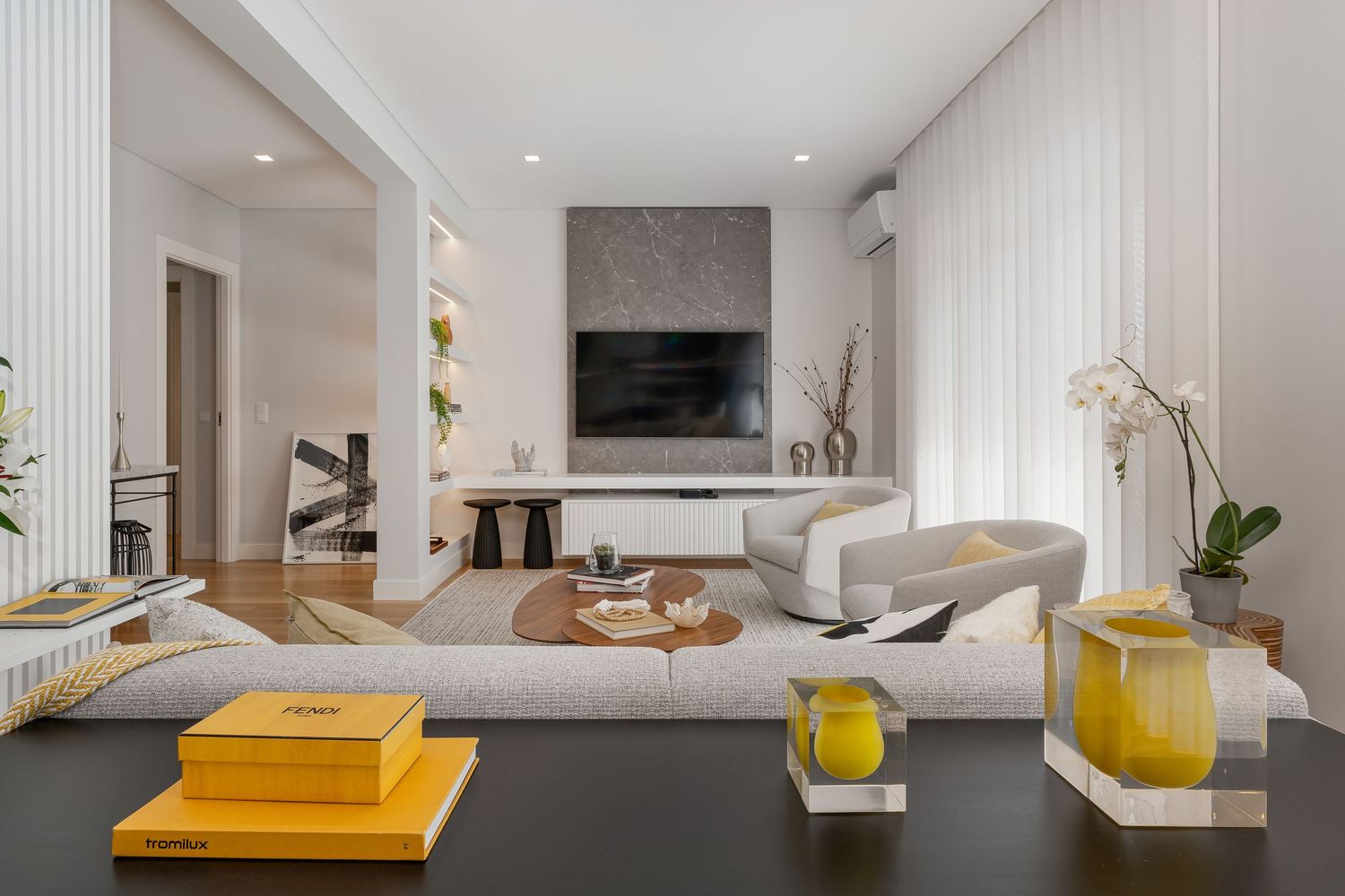 Modern living room with white walls, mounted TV, neutral furniture, and yellow accent decor on black coffee table.