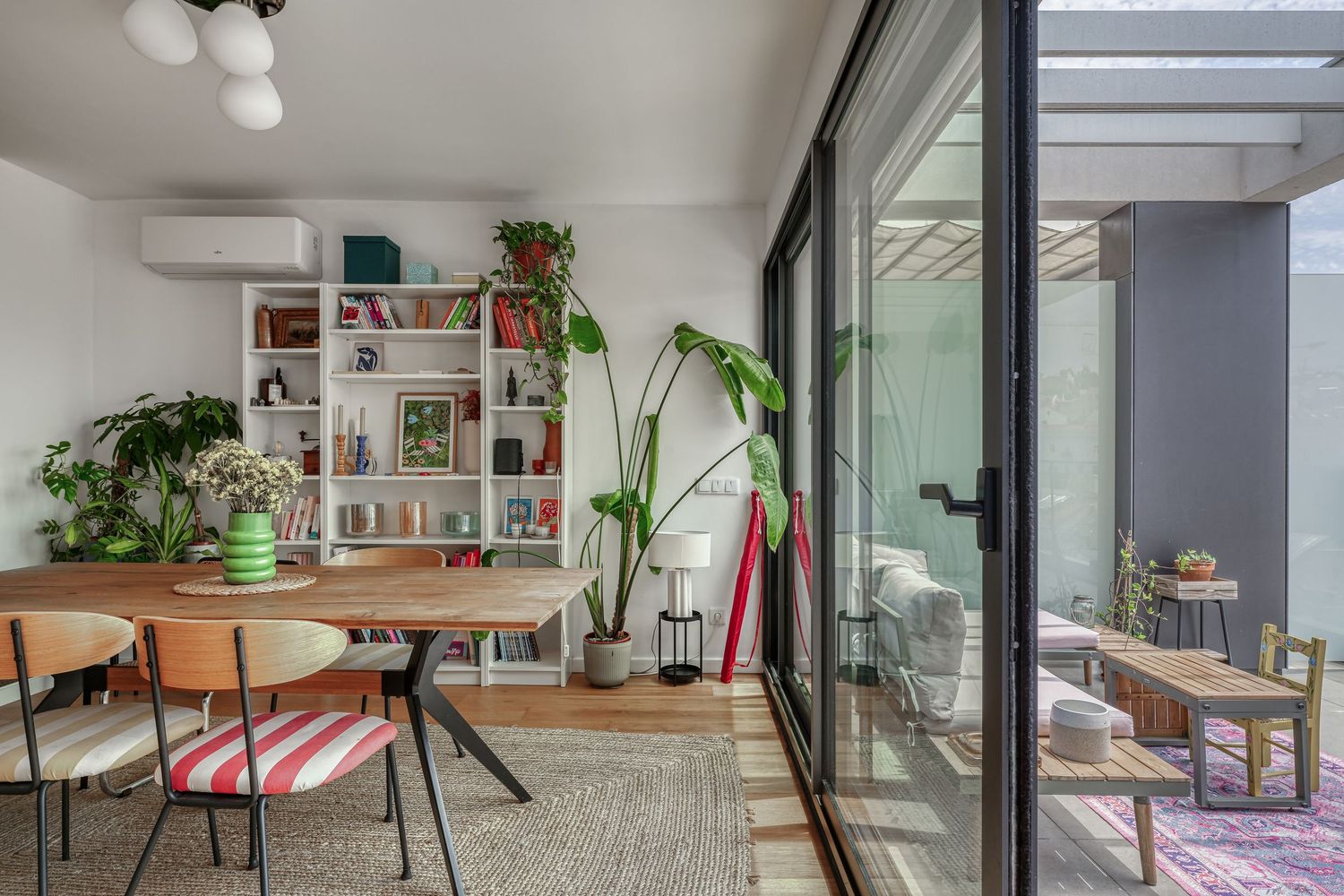 Modern dining room with wooden table, colorful chairs, built-in bookshelf, indoor plants, and glass sliding doors to outdoor patio.