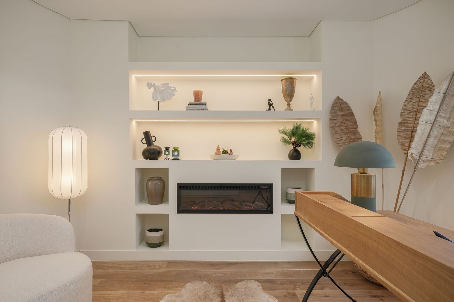 Modern living room with built-in electric fireplace, illuminated shelves, wooden desk, and neutral decor accents.