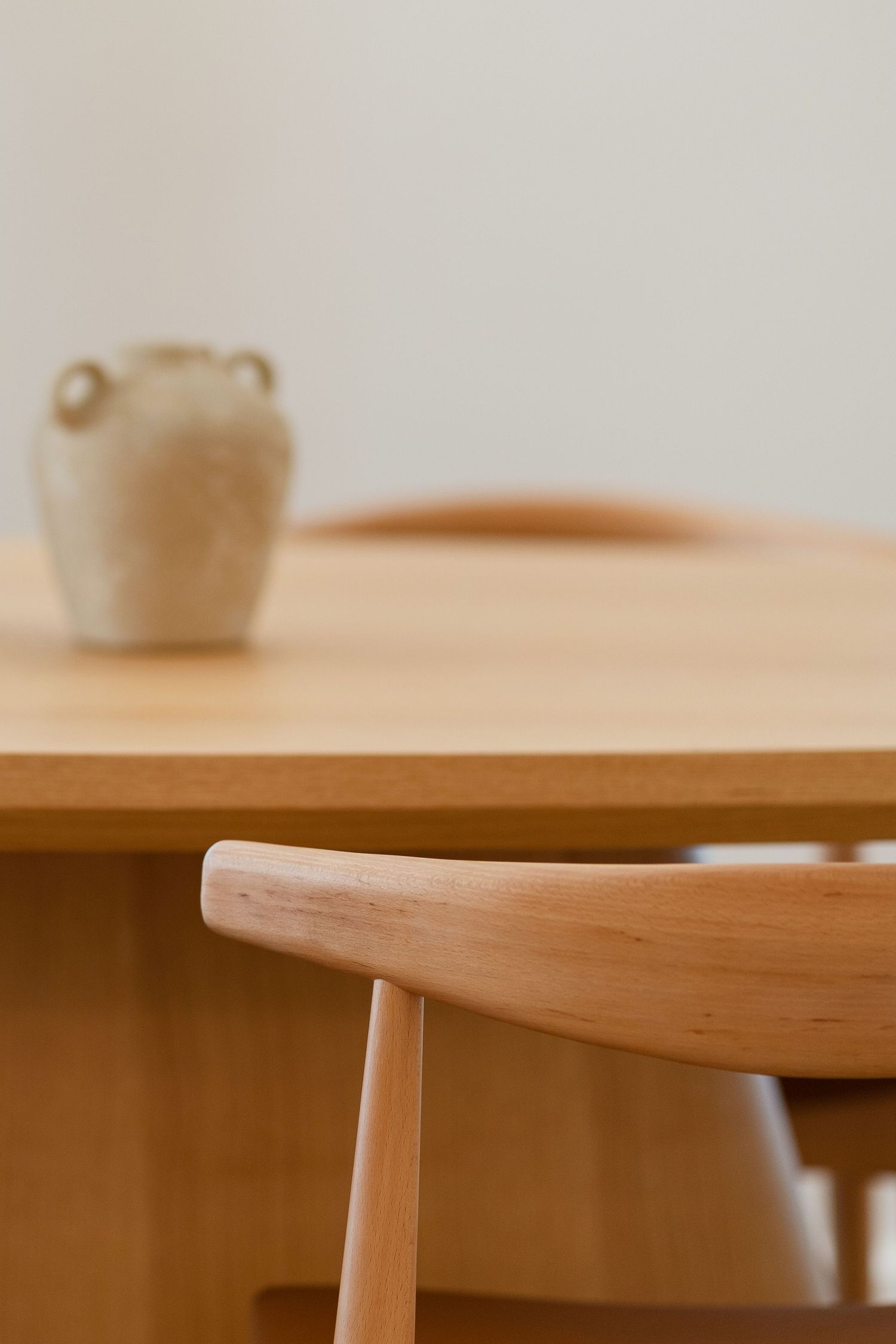 Close-up of a light wood dining table and chair with a rustic ceramic vase in the background.