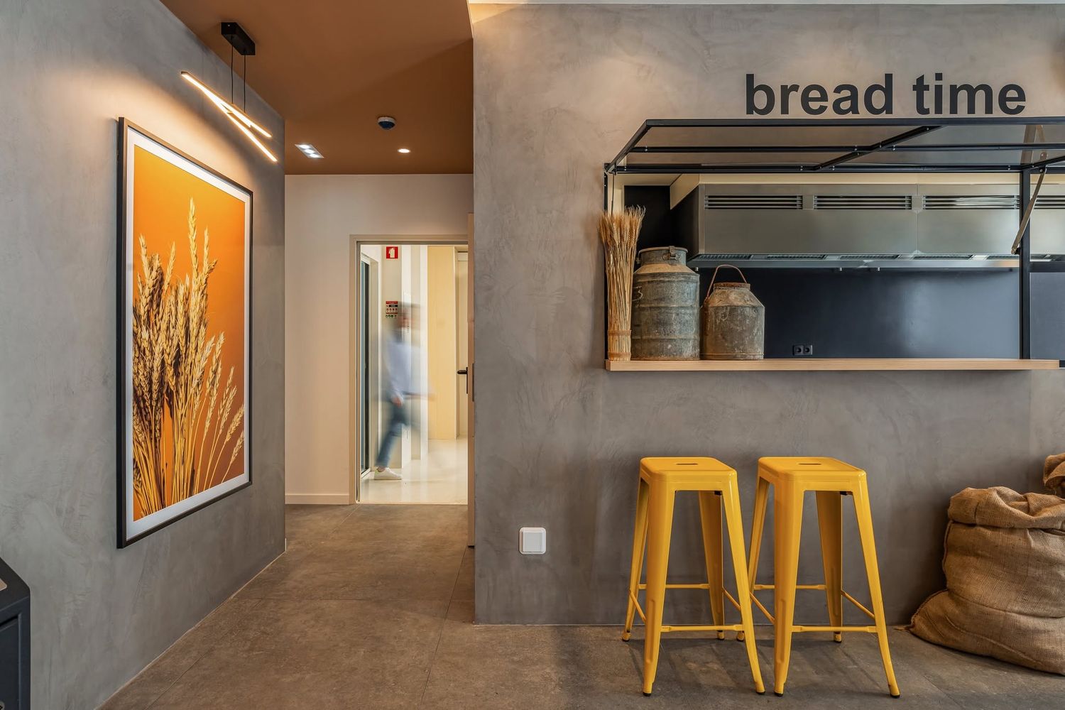 Modern cafe interior with wooden counter, yellow bar stools and minimalist concrete walls featuring bread displays.