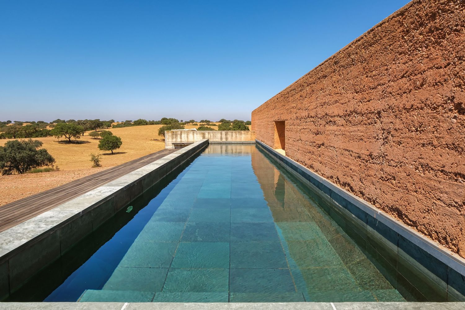 Modern infinity pool with turquoise water beside a rammed earth wall overlooking a vast golden landscape.