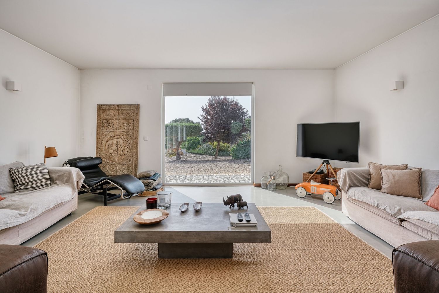 Bright modern living room with large window, gray sofas, leather chair, flat-screen TV, and a low wooden coffee table on a rug.