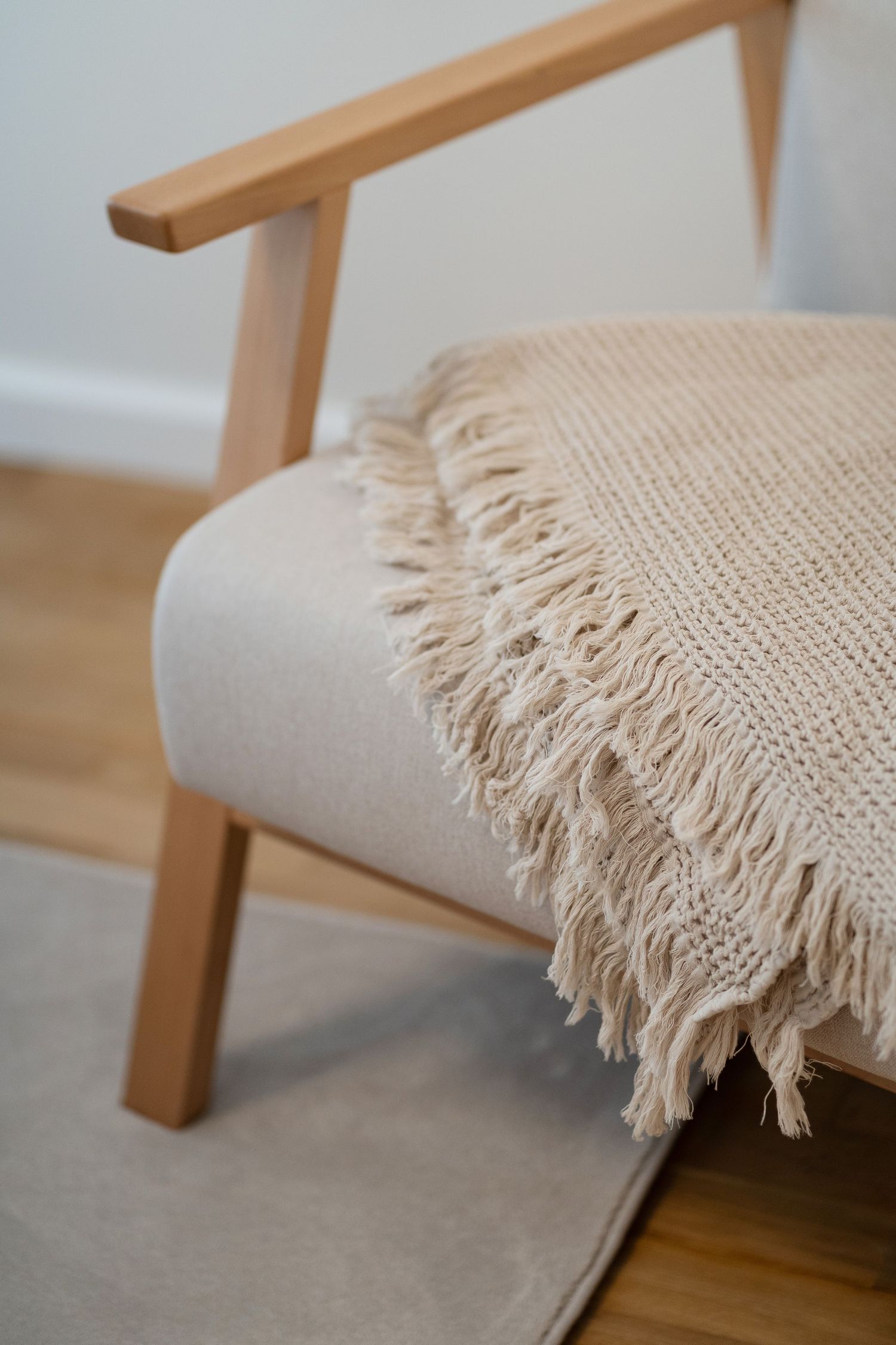 Cozy beige knit throw blanket with fringe draped over a modern wooden armchair on a light wood floor.