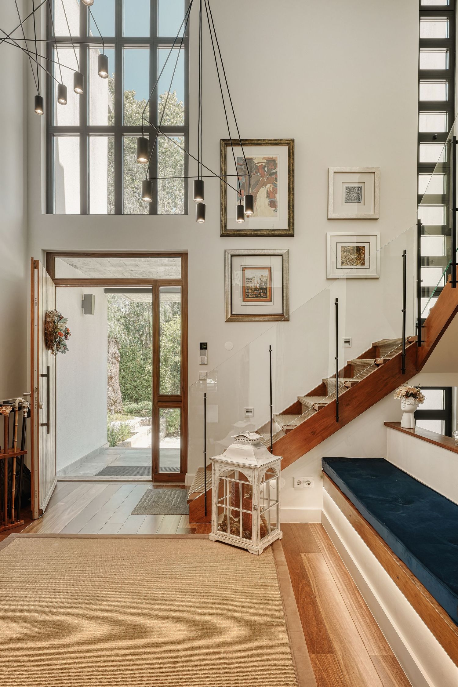 Modern home entryway with high ceilings, glass front door, wood staircase, gallery wall, and decorative lantern on hardwood floors.