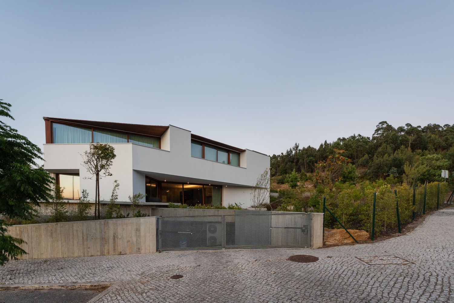 Modern luxury house with concrete walls and angular architecture set against a hillside with cobblestone driveway.