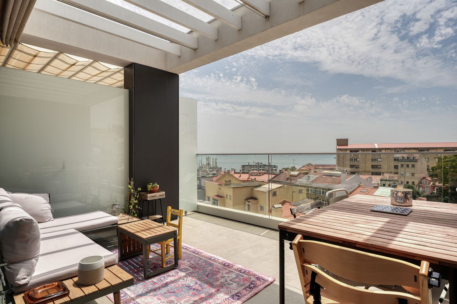 Bright rooftop terrace with lounge seating, dining table, colorful rug, and stunning ocean views over city rooftops.