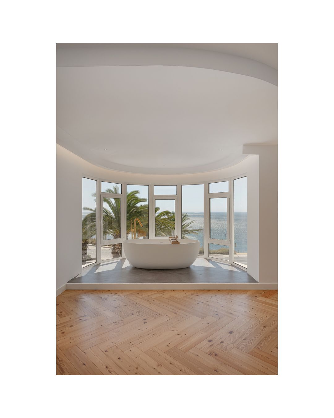 Curved window wall with panoramic ocean views and light wood herringbone flooring.