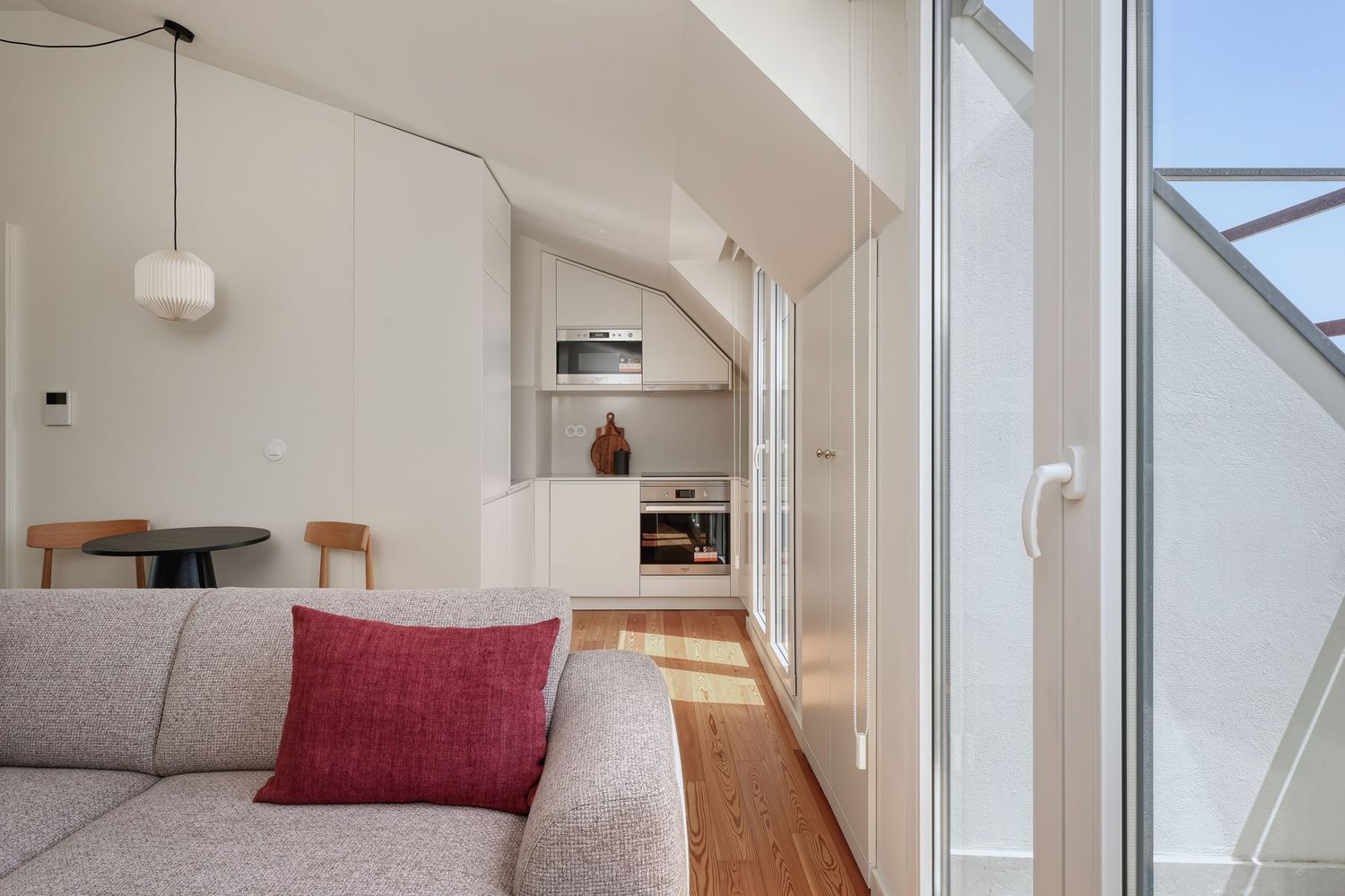 Modern studio apartment with gray sofa, red cushion, open kitchen, and glass balcony door with natural light.